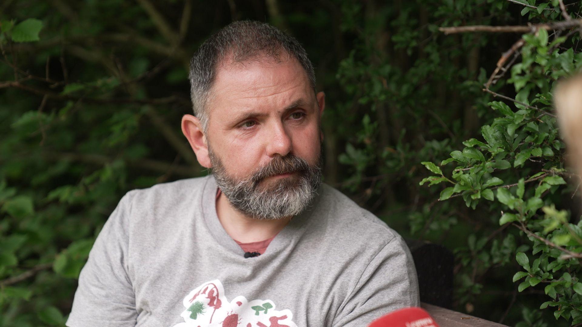 A man in a grey t-shirt looks seriously to the right of the camera. He is surrounded by green leaves.