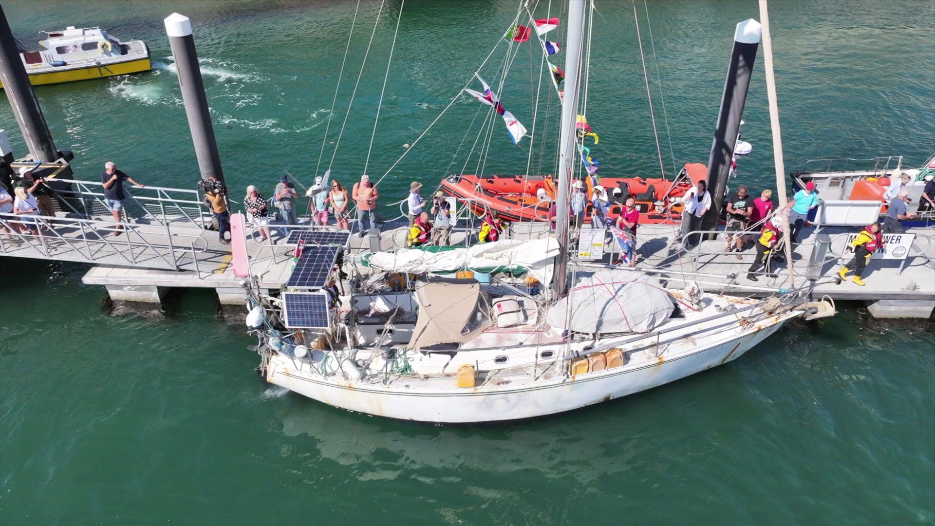 This picture shows a sailing boat moored at a dock with solar panels on the deck of the sailboat. Various pieces of equipment are visible, such as lifebuoys and ropes.
Colourful flags are hanging from the mast and there are several people on the dock.