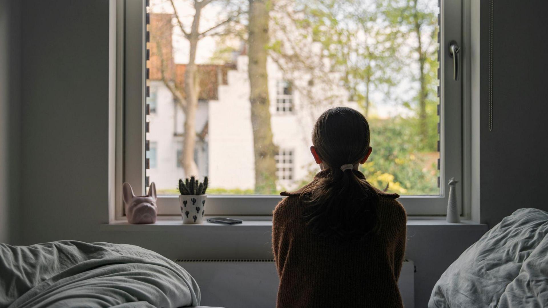 A young girl looks out of her bedroom window. The view through the window is blurry but shows a residential area.