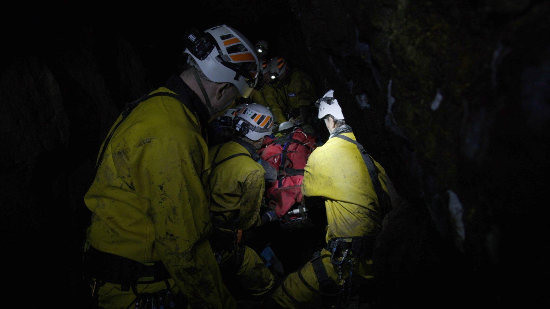 Underground rescue team COMRU in yellow overalls surround a red-blanketed stretcher inside a cave