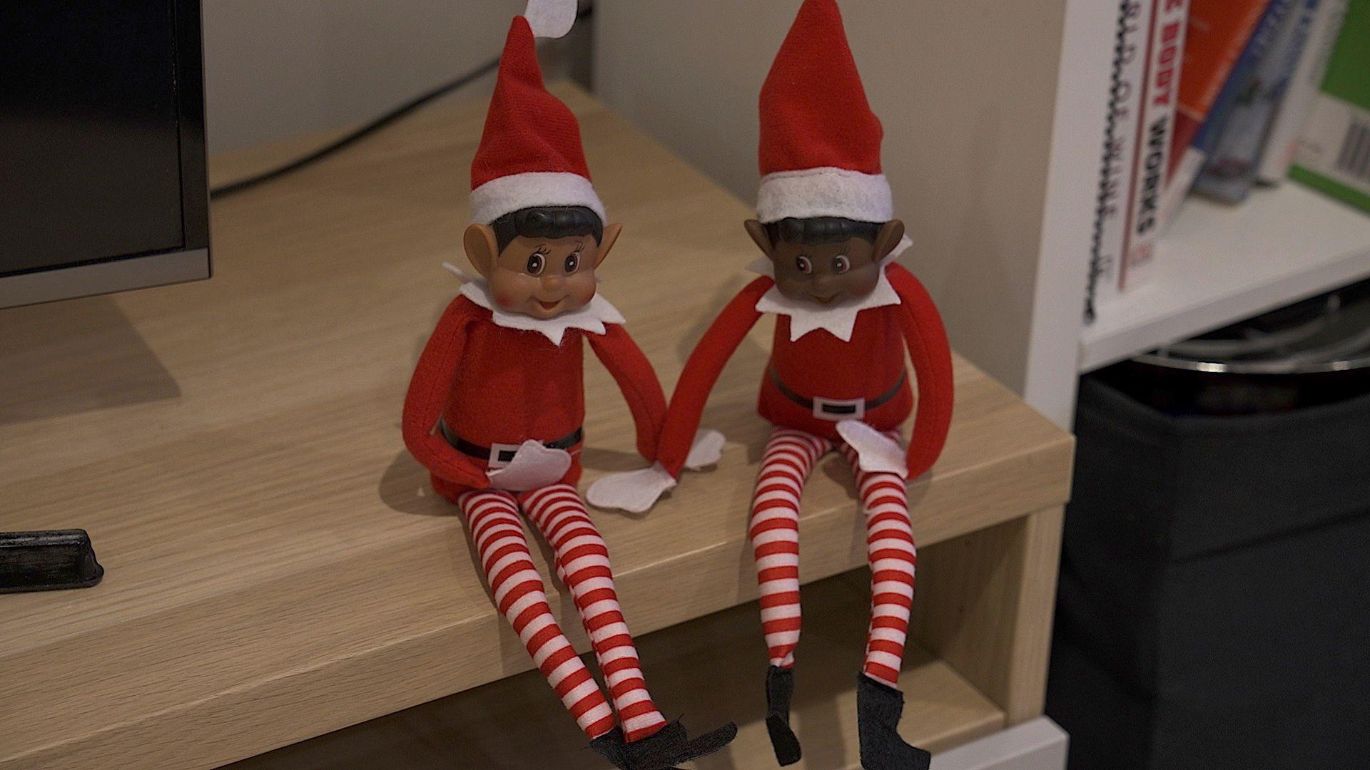 Two toy elves with red and white stripy tights sit on a wooden television stand with part of the TV visible.