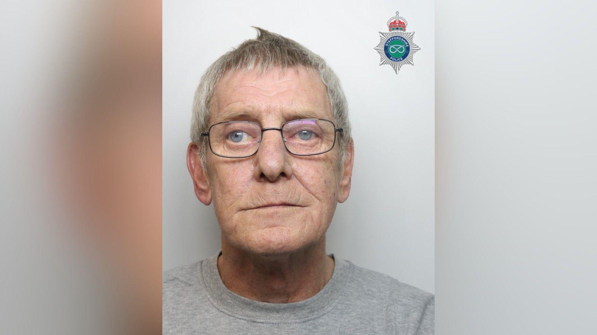 A mugshot of an elderly man with short grey hair and glasses. He is wearing a grey top.