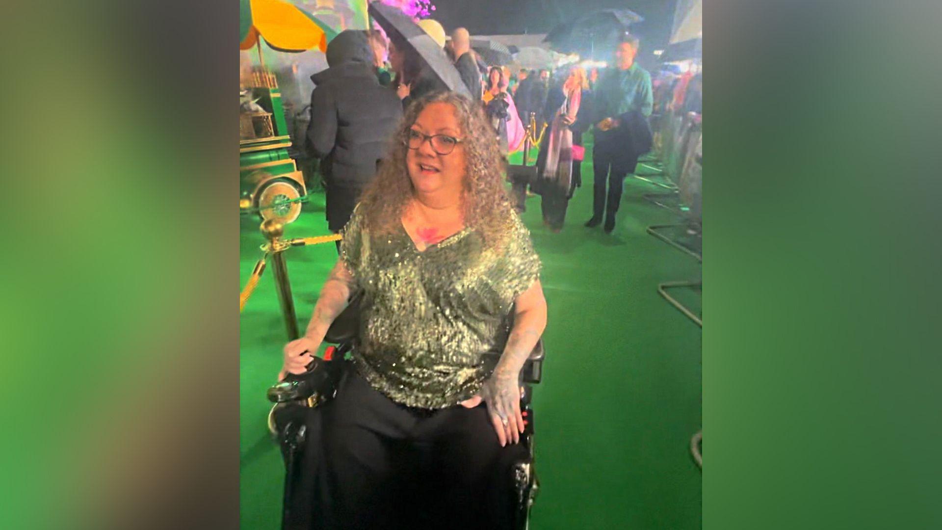 Chantelle Nassari sat in a wheelchair. She is wearing a green sparkly top and has blonde curly hair and is wearing glasses. She is at the premiere for the film and there are people standing behind her.