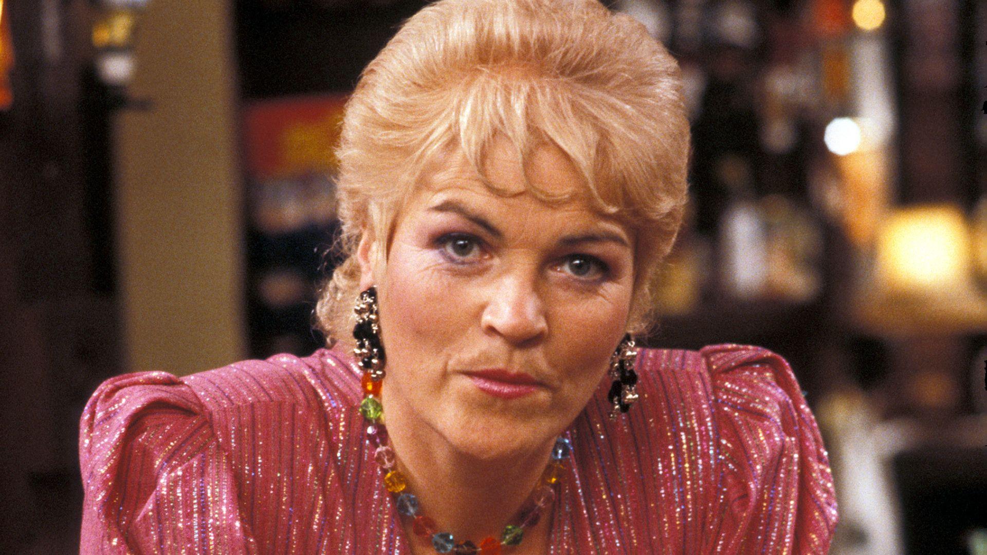 portrait of pat butcher, a woman with blonde bouffant hair, a shiny pink dress with shoulder pads, large dangling earrings and a necklace of multiple coloured beads