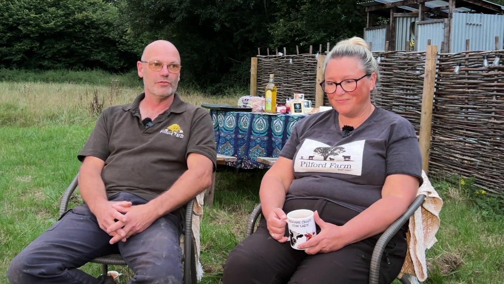 a man with a shaved head and a blonde woman sit in chairs outside in front of a wicket fence and small table.