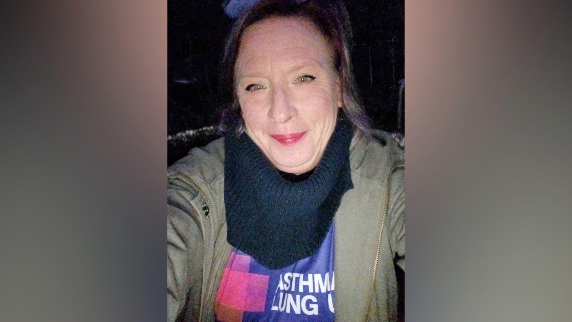 Chloe Slasberg, wearing a green jacket with black scarf, on top of a purple T-shirt with Asthma and Lung UK branding on. Chloe is smiling at the camera.