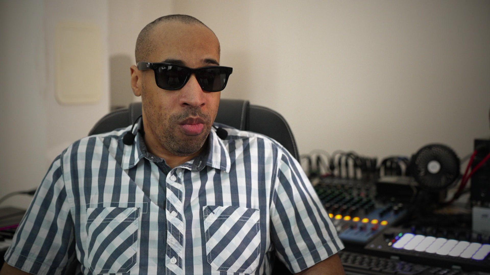 Andre Louis sitting in his house wearing a black and white striped shirt and with sunglasses on
