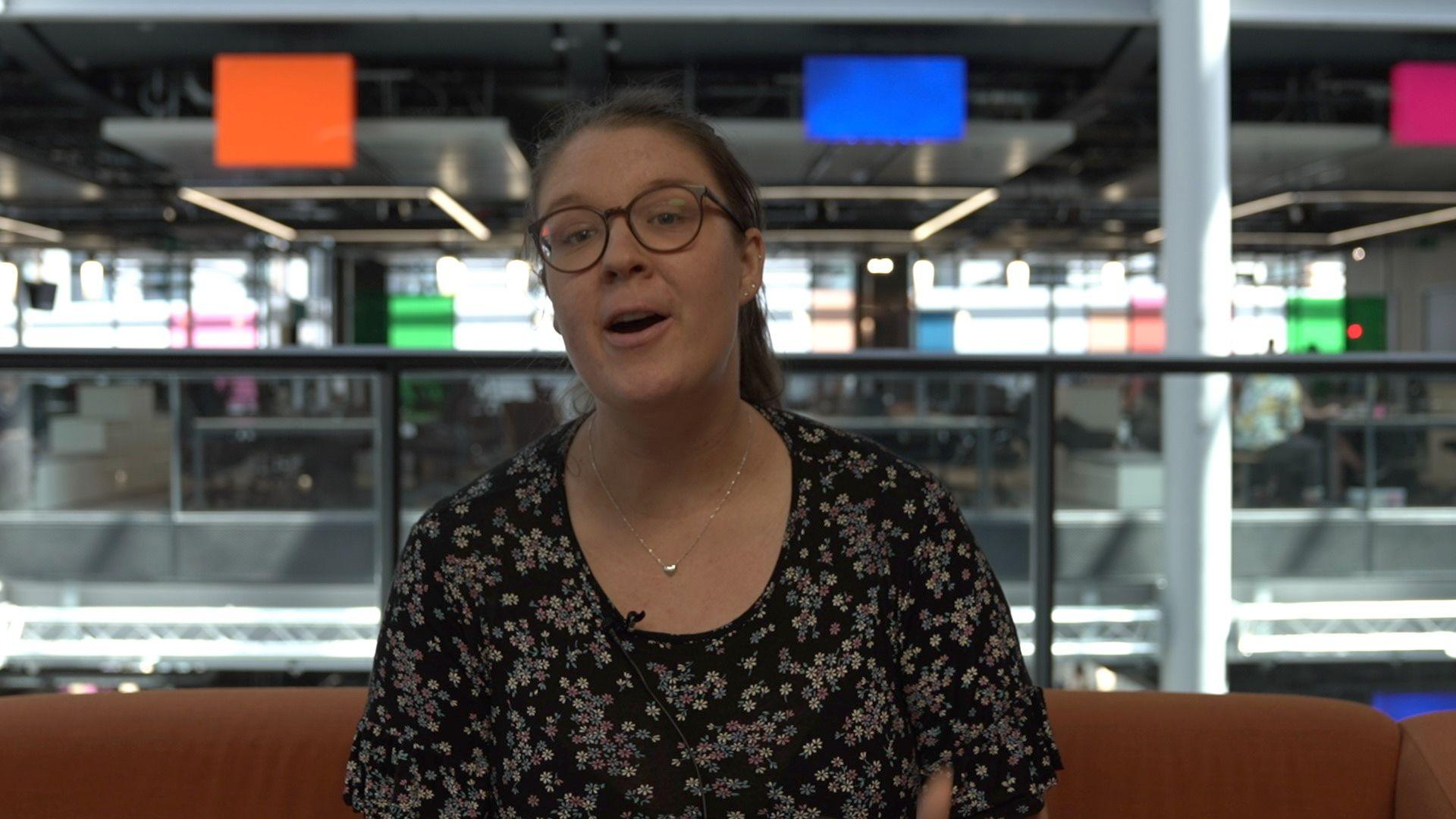 Catriona Aitken - who has glasses and tied-back brown hair and is wearing a black floral dress - sat on an orange sofa, with the colourful panels of the ý Wales office behind her