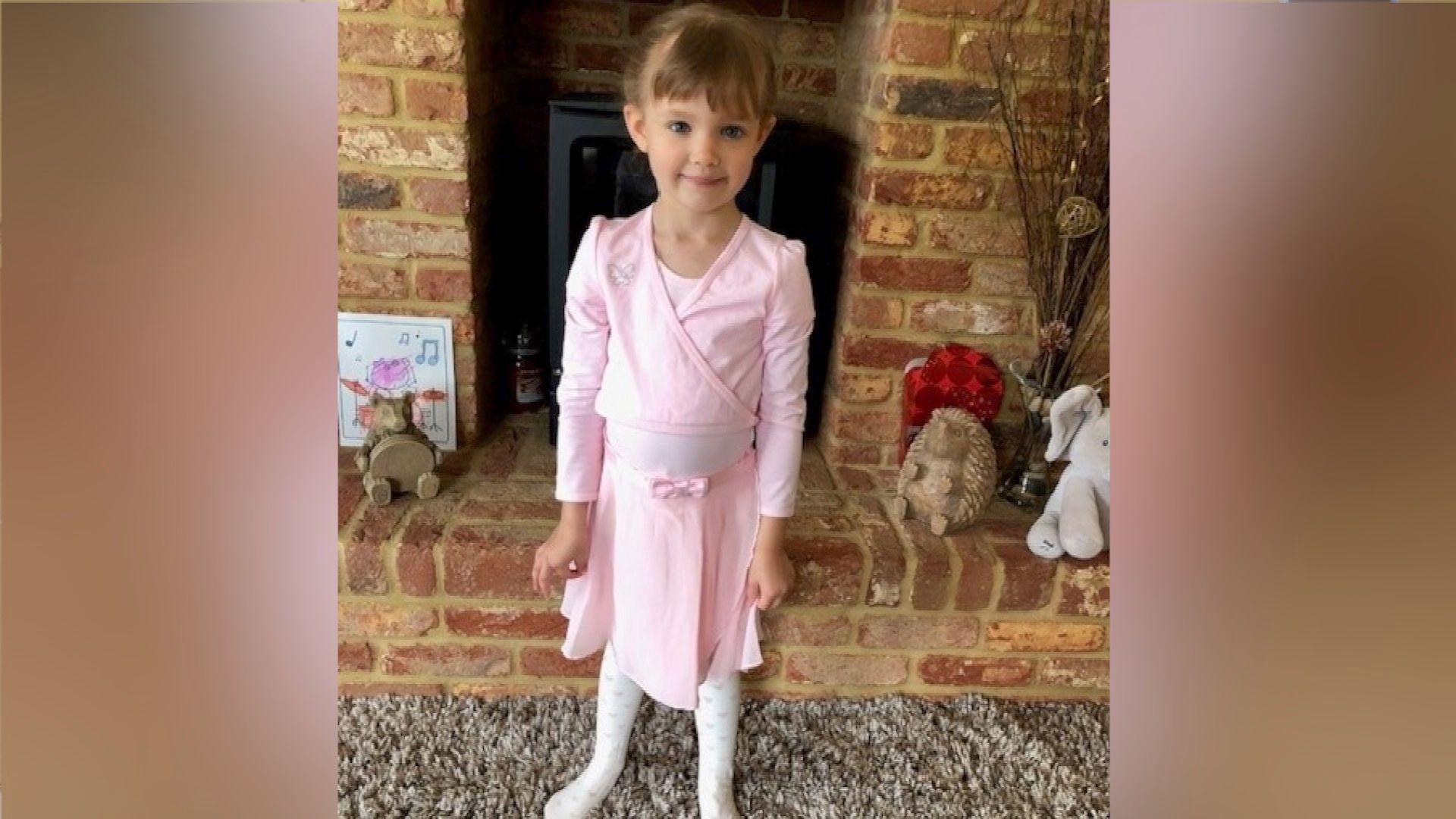 Jessica in a pink ballet outfit, with brown hair, her hair tied back, standing in a living room in front of a brick fireplace.
