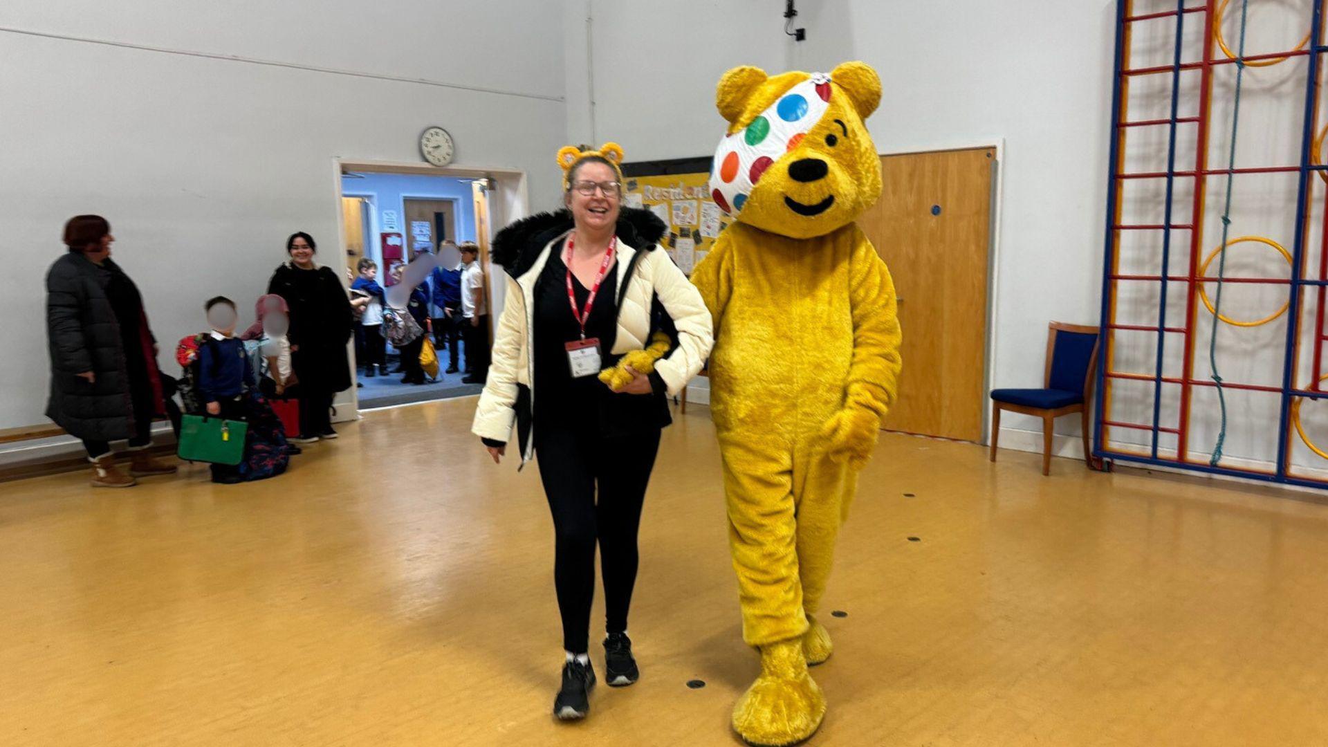 A woman in a white puffer jacket standing next to a large yellow bear mascot inside a school hall