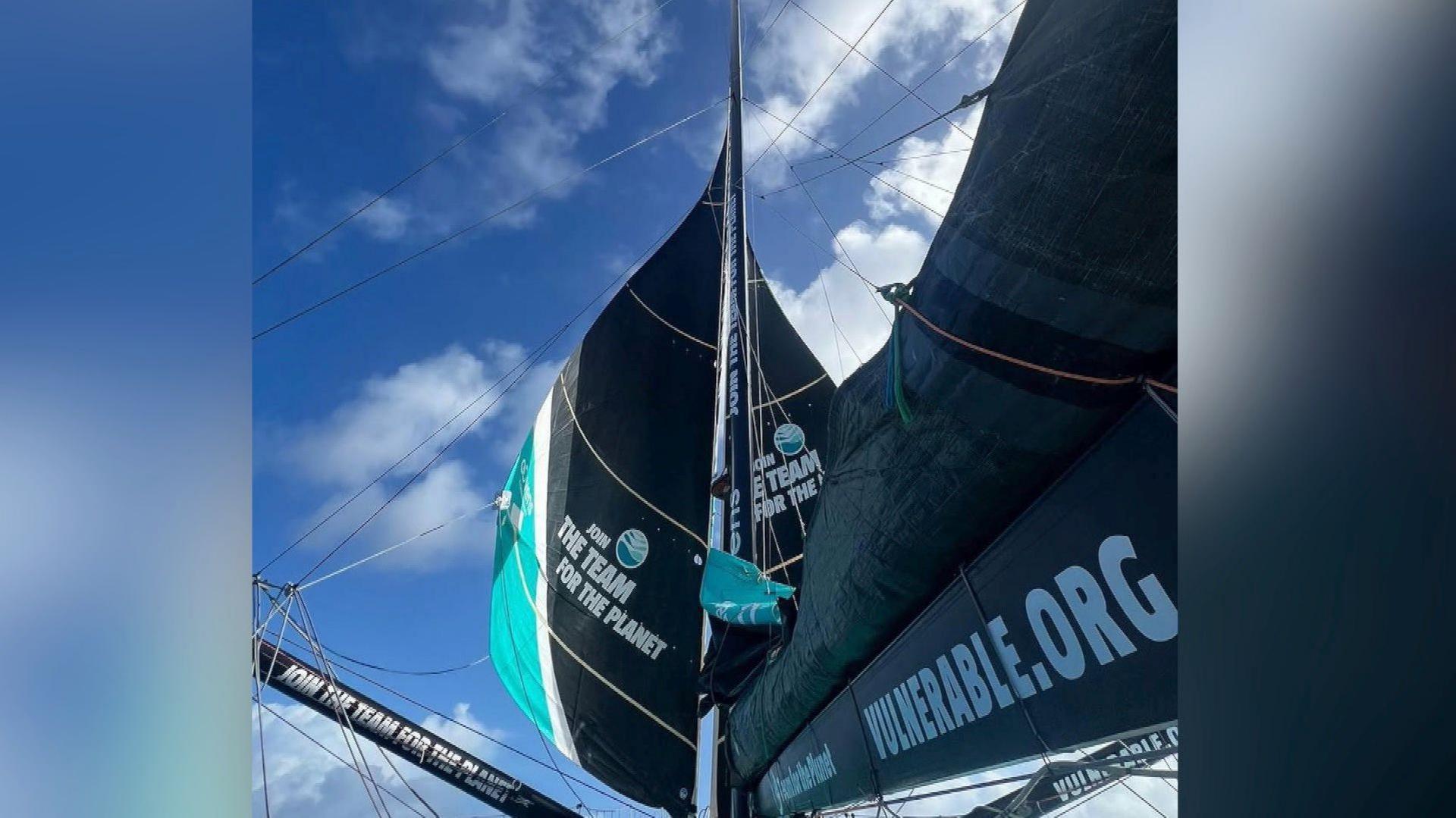 Two jibs on either side of the mast with a mainsail folded up and a number of written logos with blue sky above.