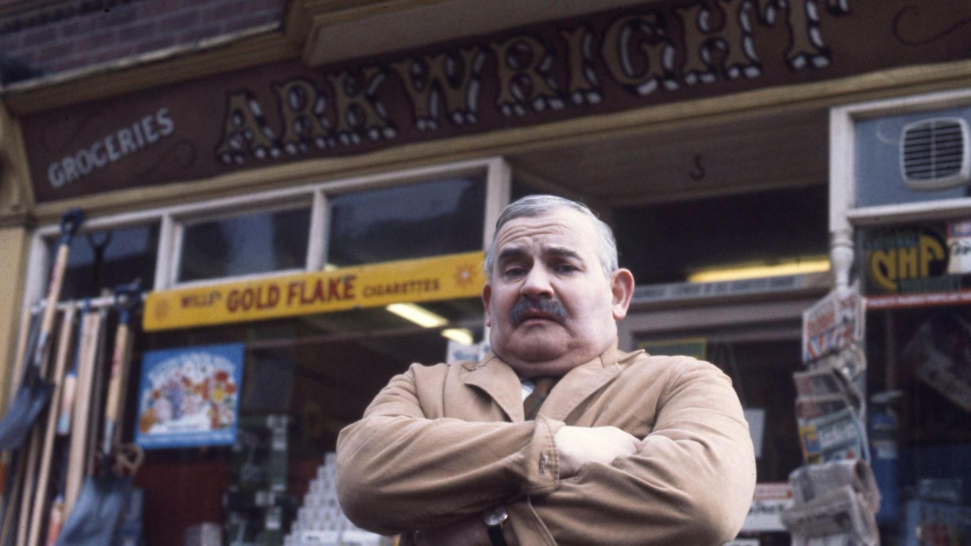 Ronnie Barker stands with his arms crossed in front of Arkwright's. He has a moustache and short grey hair.