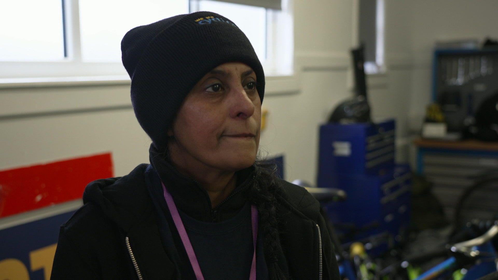 An Asian woman, wearing a black beanie hat and black coat, looks to the right of the camera as she stands in a bike room