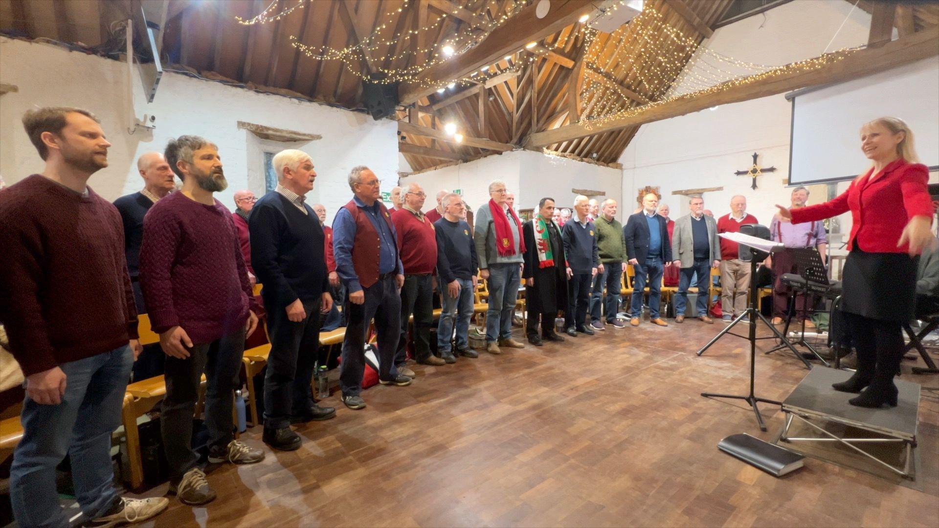 The Welsh Male Voice Choir in Oxford singing in a room. A woman conducts them. There are lights on the ceiling and a crucifix.