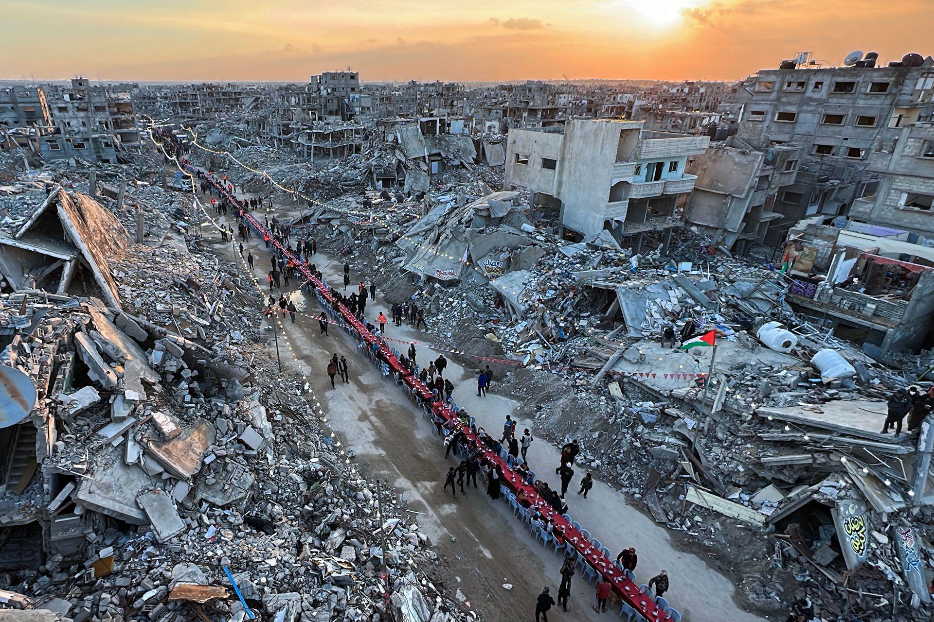 A wide aerial view of Rafah shows mass buildups of rubble and devastation around a long red table running down a street where residents break their fast on the first day of Ramadan in March 2025.