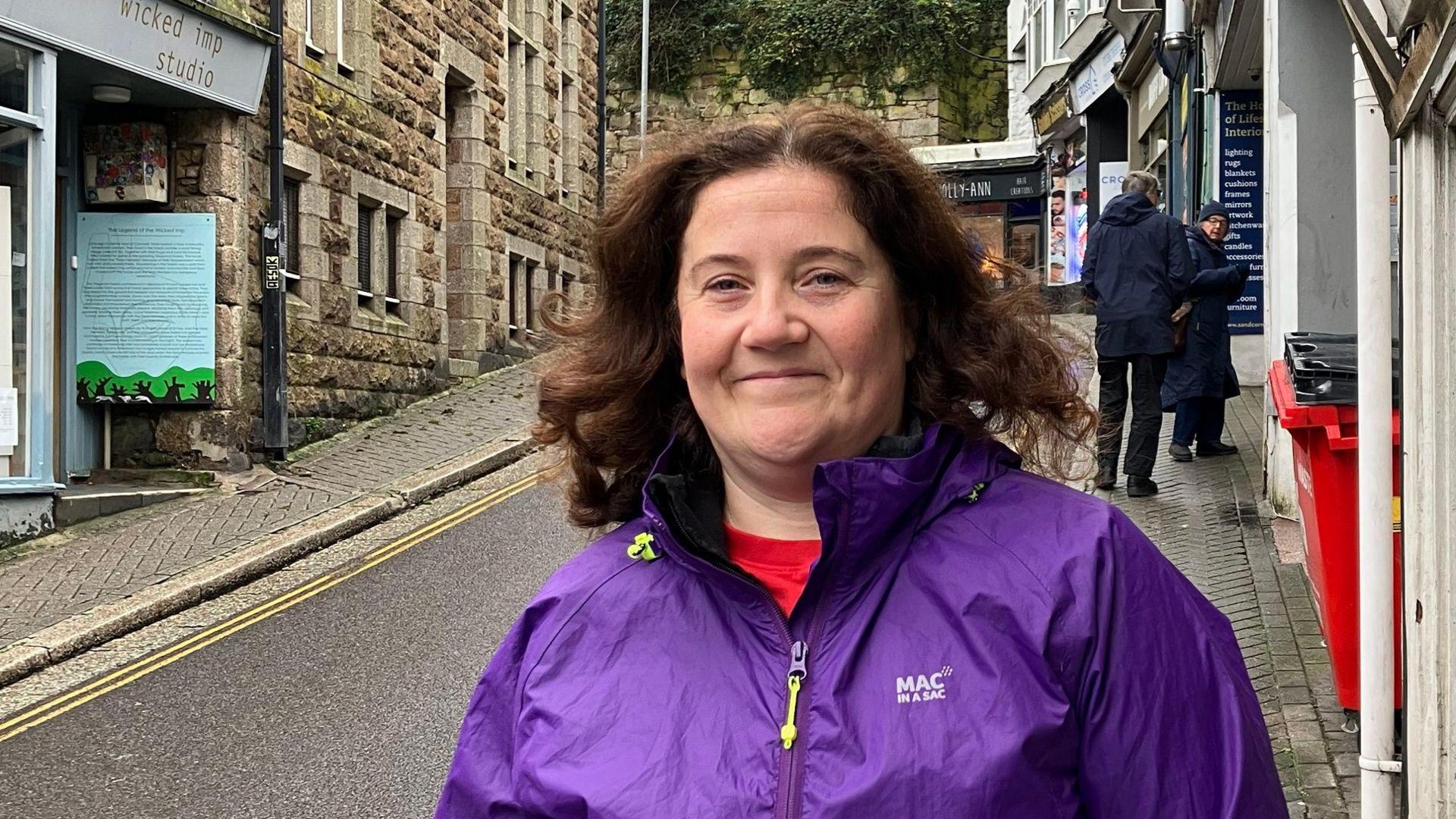 Natalie Preston is smiling as she stands on the pavement of the narrow street which leads up hill. She is wearing a purple anorak and has shoulder-length brown curly hair. There are small shops lining the street.