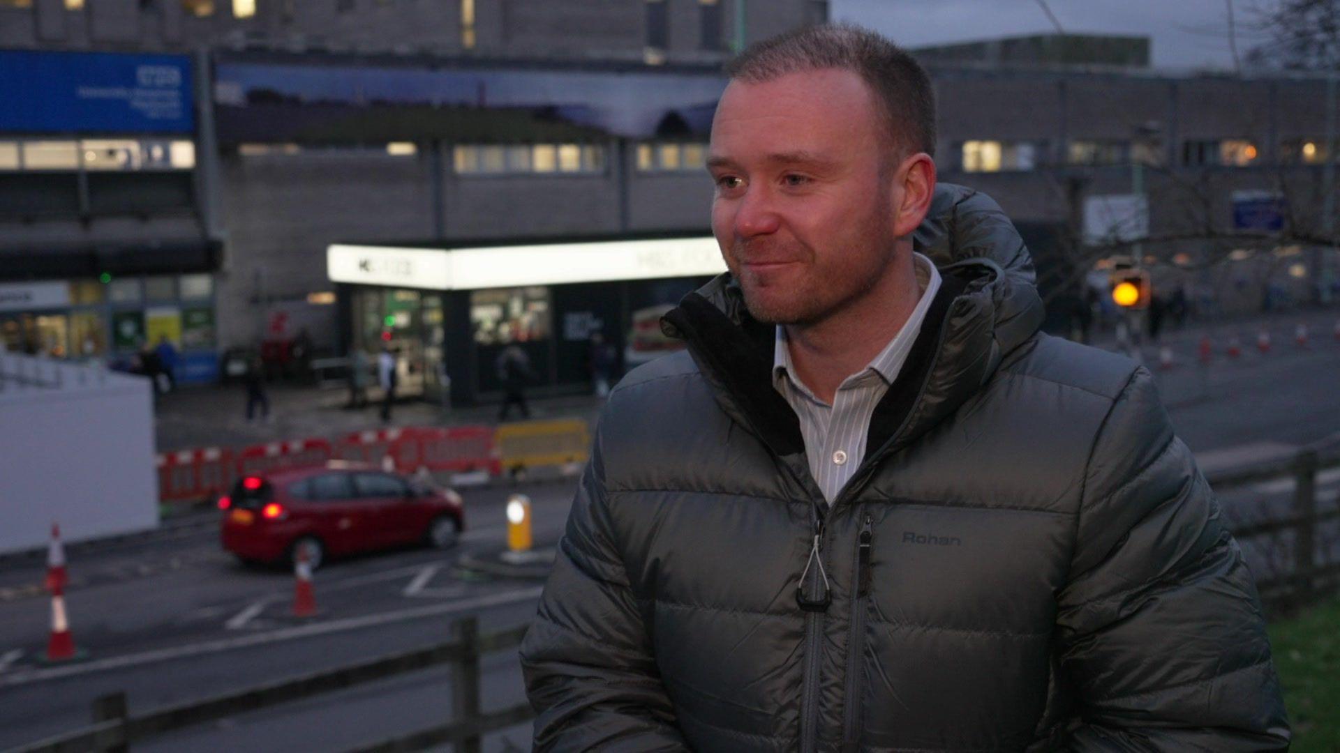 A picture taken from an interview with Fred Thomas MP outside of Derriford Hospital, Plymouth. He is wearing a light blue and white striped shirt with a black coat on top. He has a short beard and short black hair.