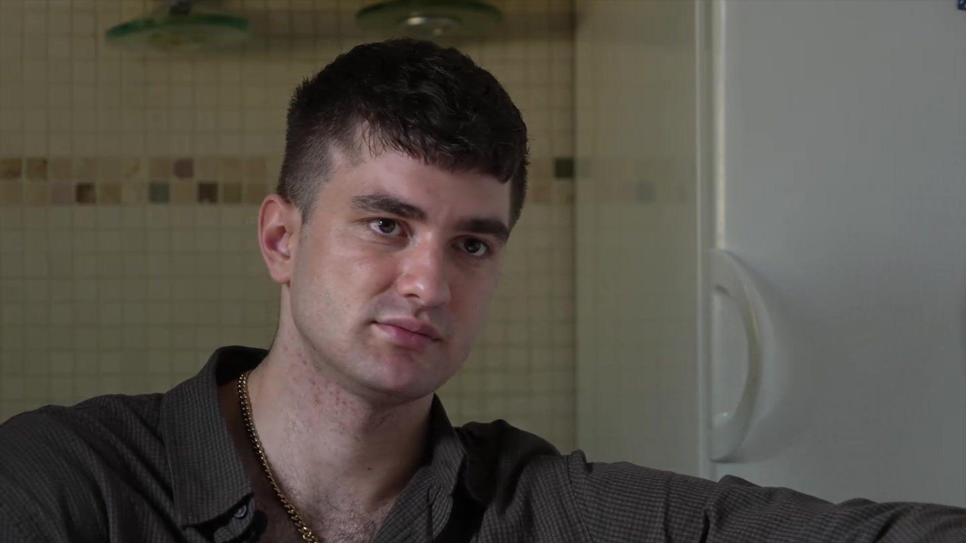A man wearing a brown shirt. He is sat in front of a tiled wall with a white cupboard in the background.