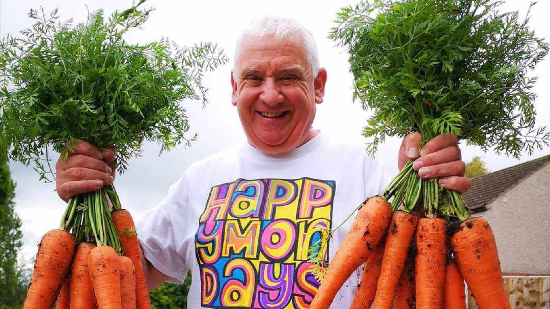 A man in a white t-shirt holds two huge bunches of over-sized carrots. He has short white hair and his t-shirt has a colourful square logo which says 'Happy Mondays' on it.