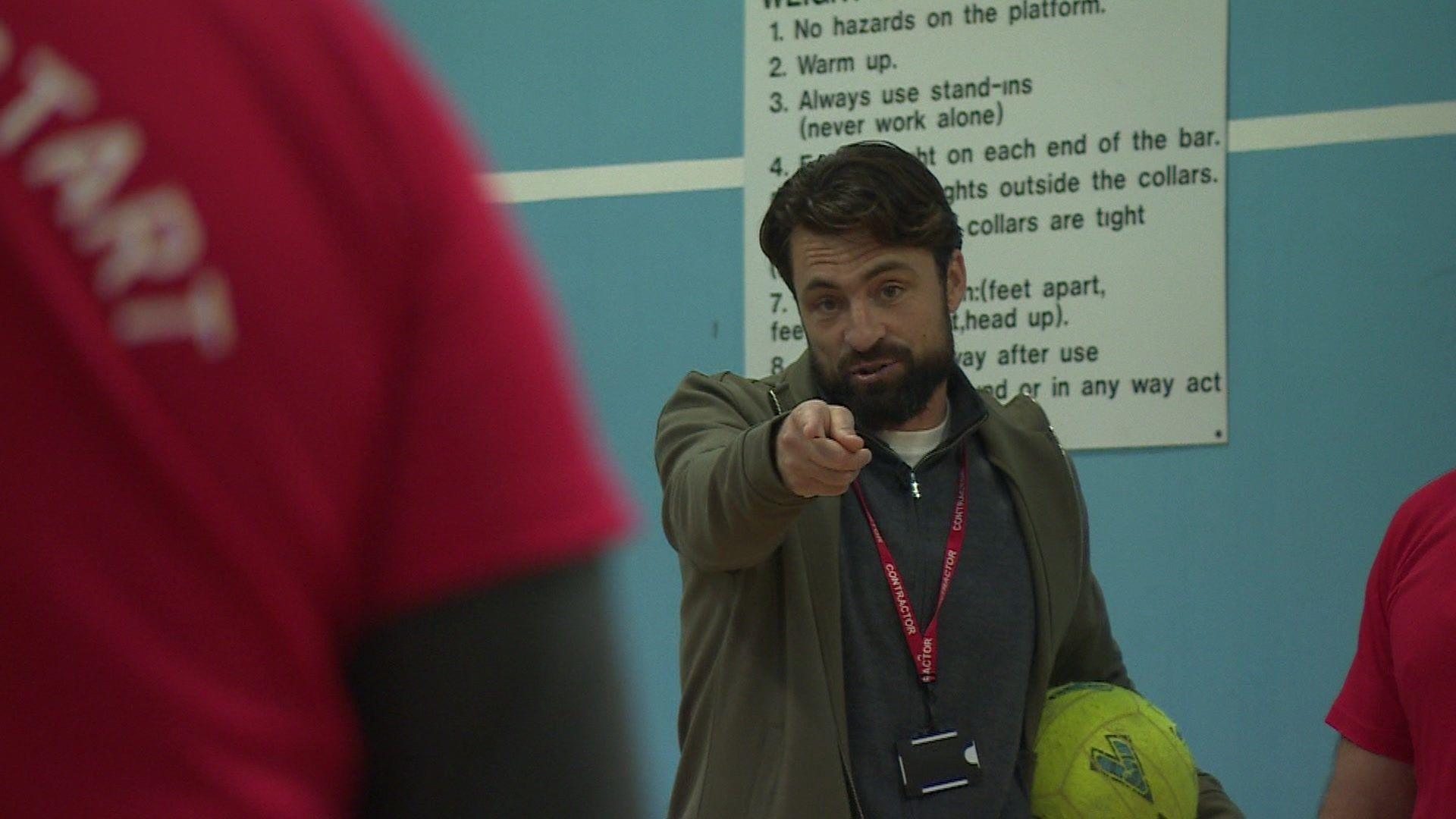 Russell Martin wearing a green jacket , holding a yellow football while pointing. Unidentifiable men in red t-shirts can be seen standing around him.