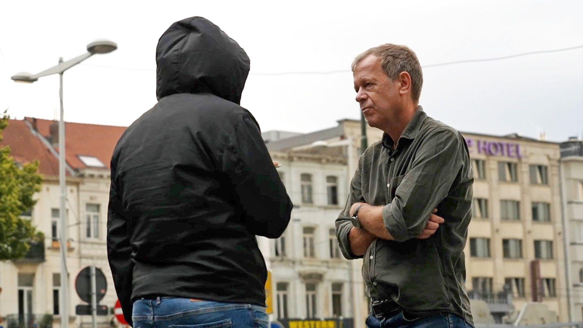Man in black coat with hood up, with his back to us stands opposite Andrew Harding who is wearing a green shirt and has his arms crossed.