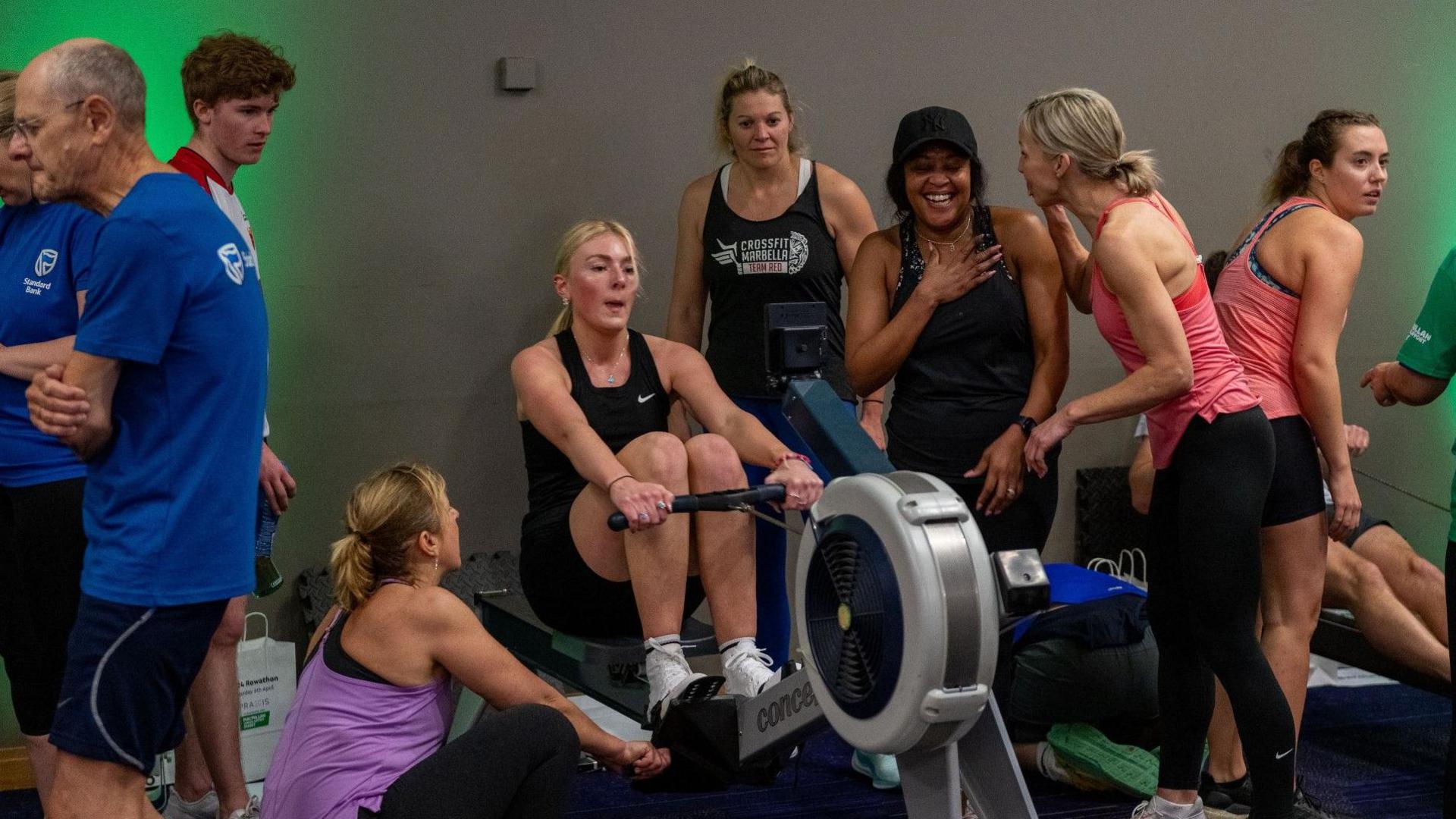 People taking part in an indoor rowing challenge. A woman is on the rowing machine. People around her are smiling and supporting her. 