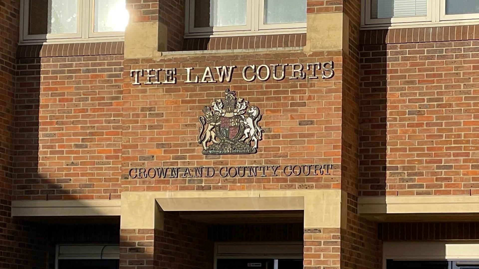 A court logo and a sign which reads 'The Law Courts Crown and County Court'. It's attached to a light brown brick wall above the entrance 