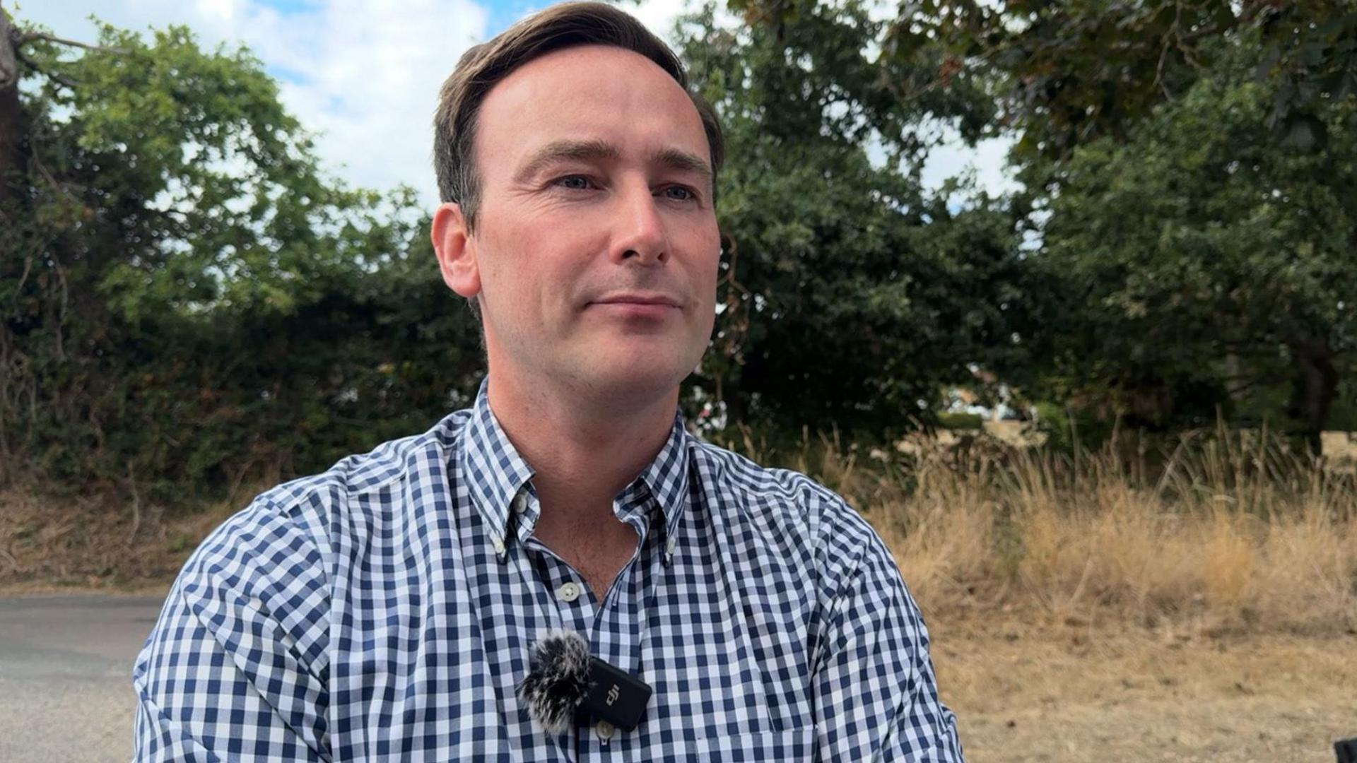 Tom Hayes MP, standing on a rural road near Throop. He is wearing a blue and white checked shirt. A hedge and field with trees can be seen in the background