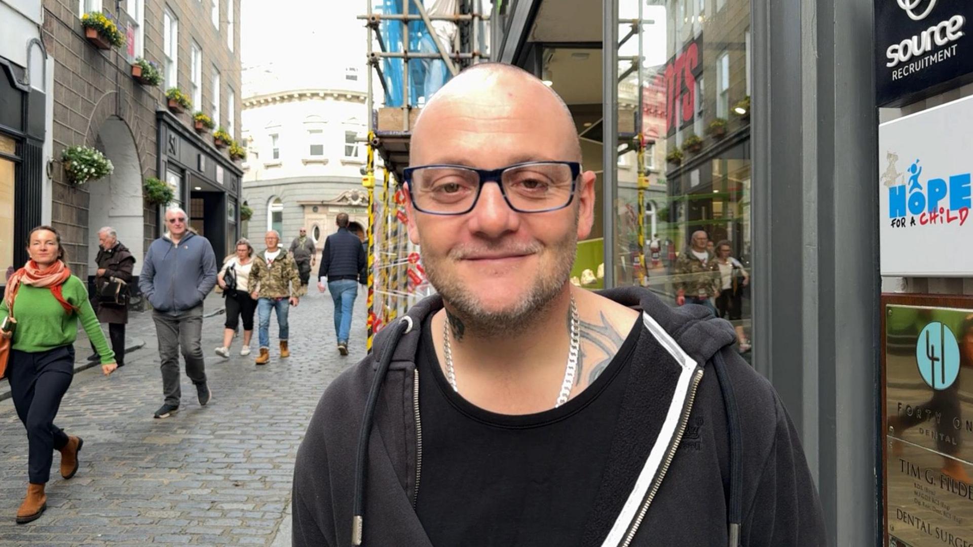 A man in his late 30s wearing a black hoodie and square glasses. He is bald but has a beard.