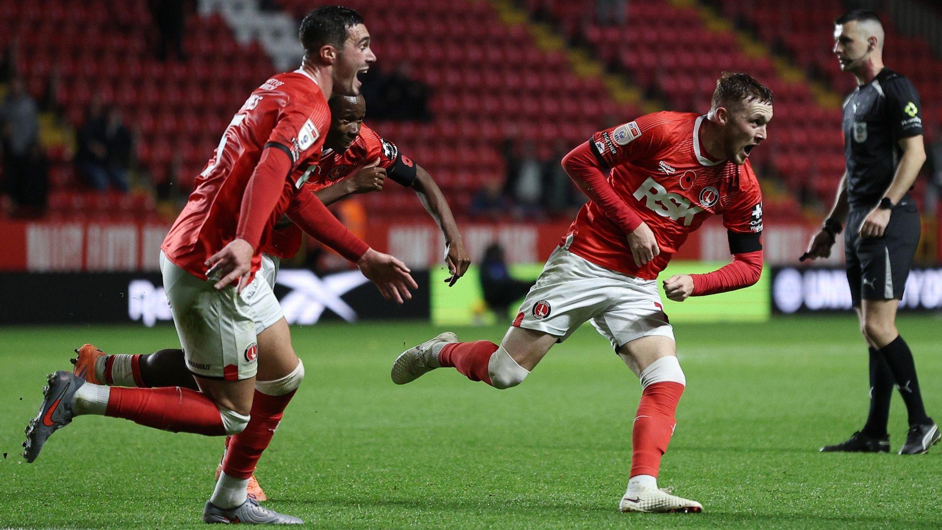 Three Charlton players wearing red shirts, white shorts and red socks run together with mouths agape and joyful expressions on their faces.