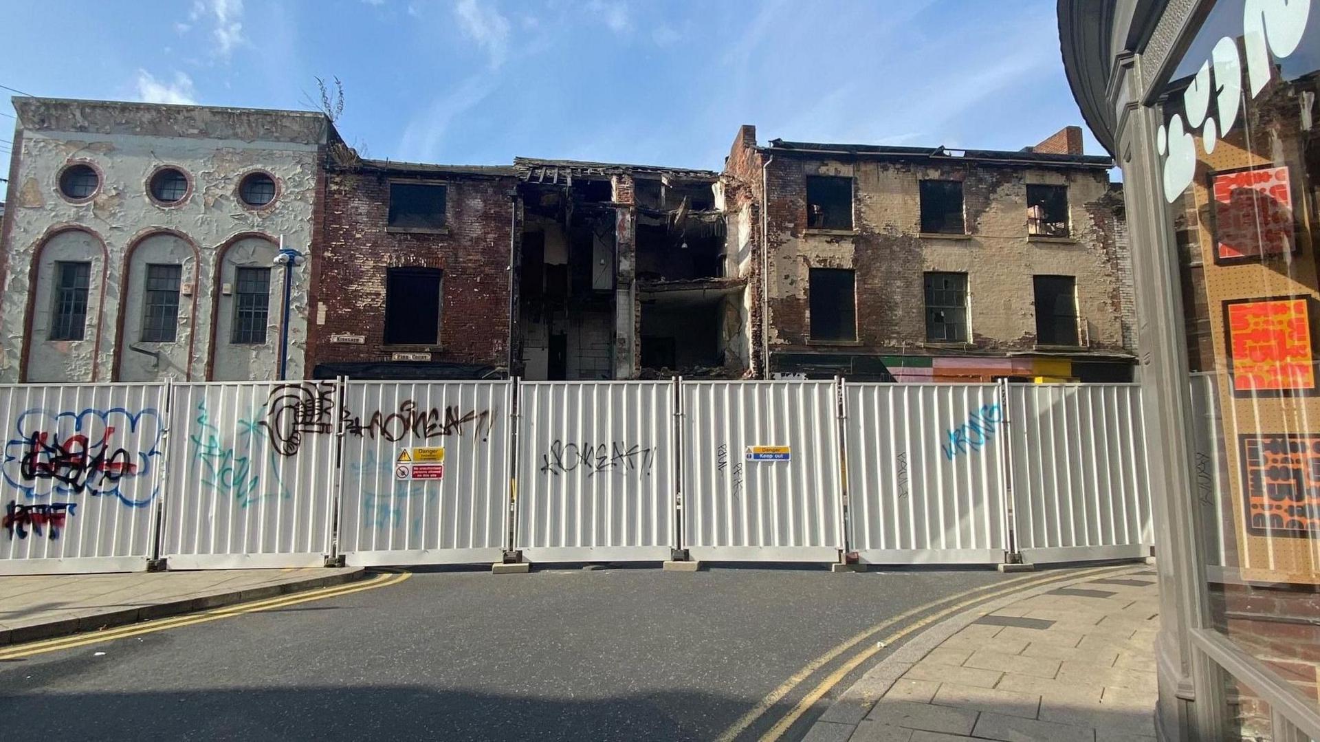 A picture showing a row of derelict buildings on a street with safety barriers cordoning them off. The barriers have graffiti writing on them. 