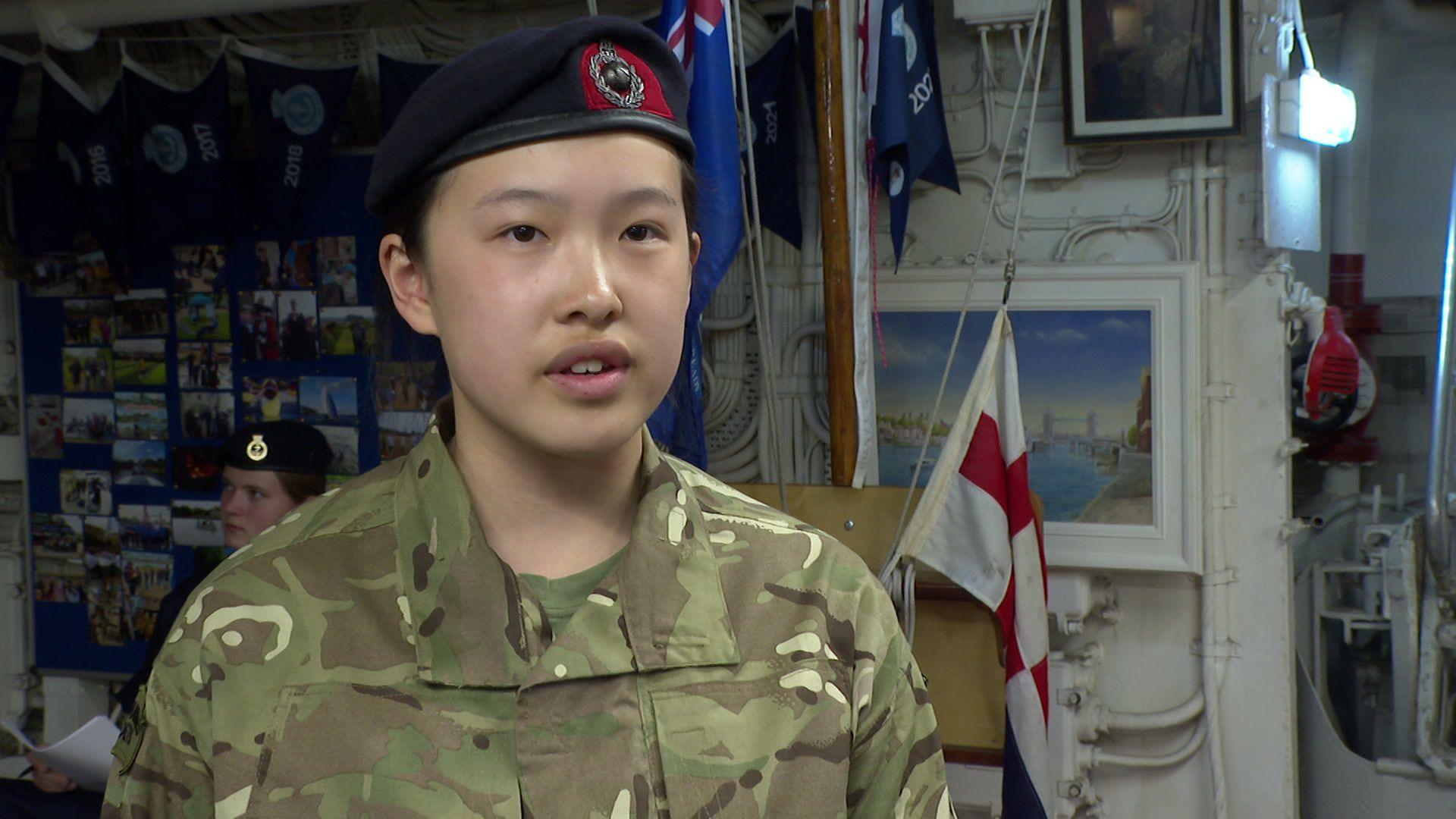 A teenage girl in a military uniforms stands in front of a flagpole.