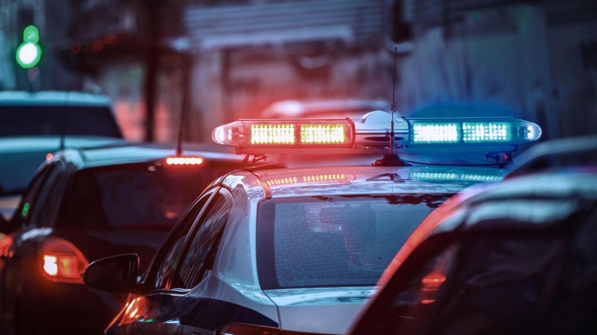 The image shows police vehicles with flashing blue and red lights queueing behind other cars in the dark.