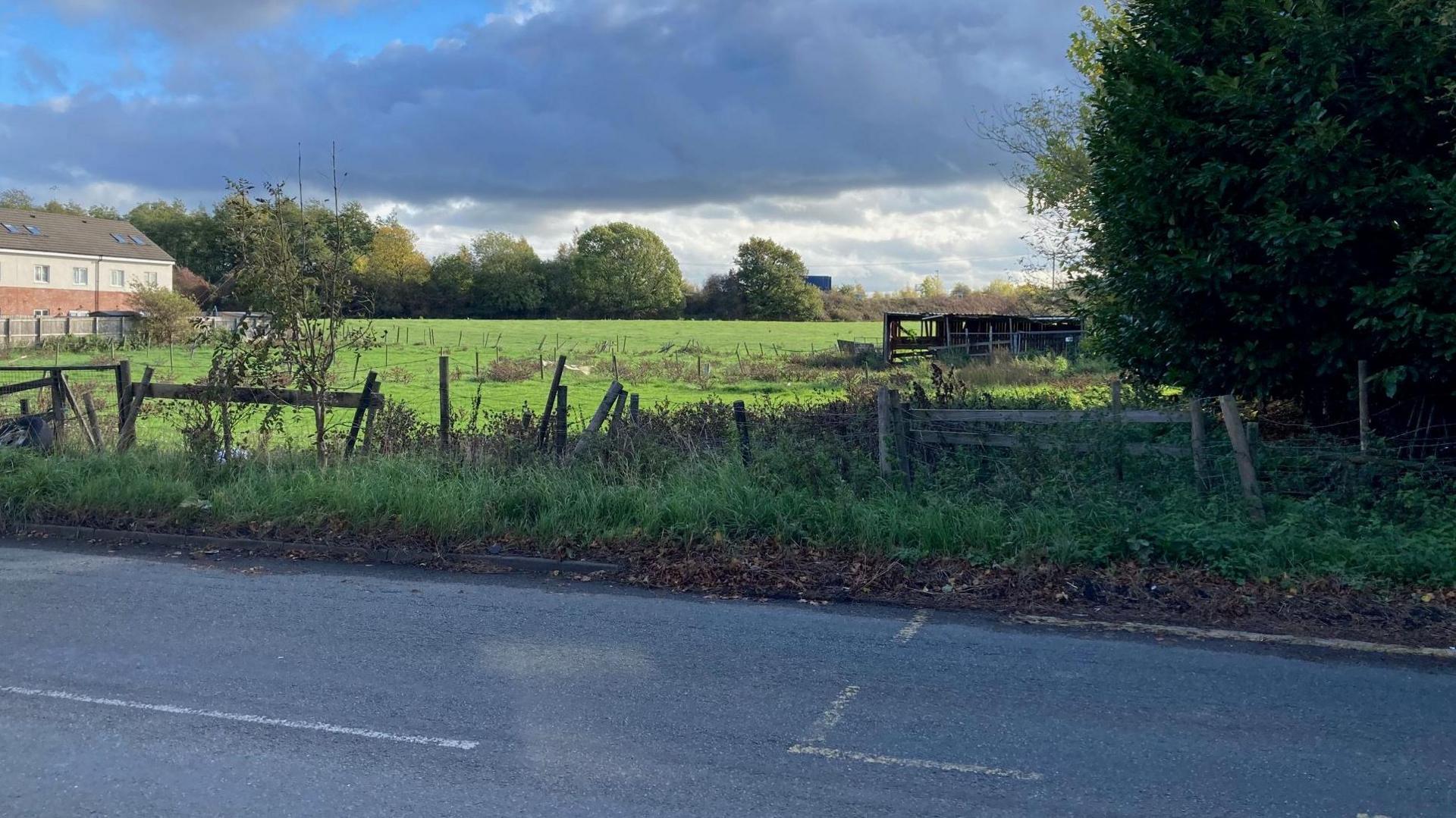 A field of grass with derelict agricultural buildings taken from the road with a number of new homes to the far left of the picture