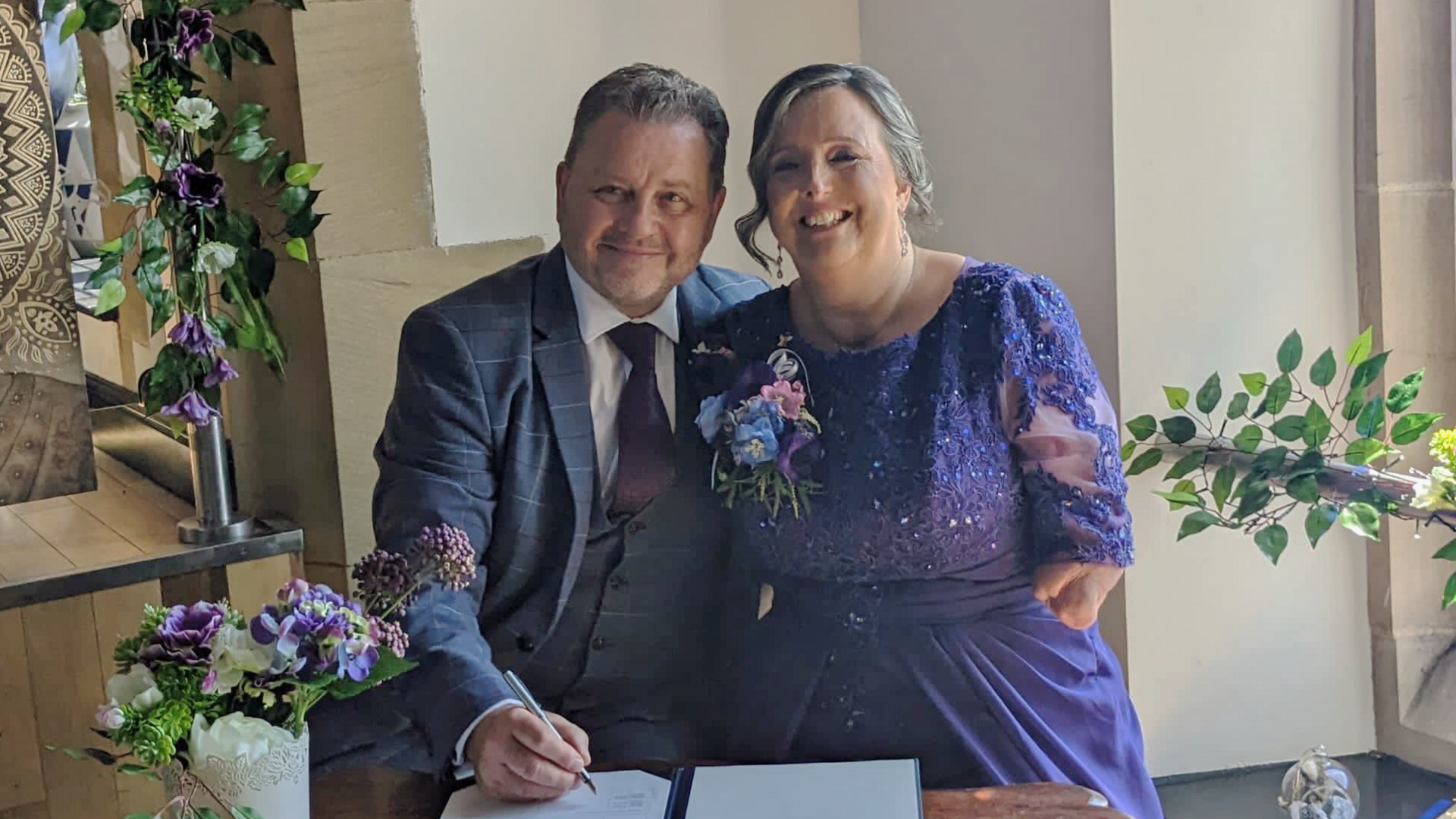 David and Allison Friday on their wedding day. They are standing smiling broadly while signing official papers. David has short dark hair and is wearing a suit. Allison is wearing a purple dress and has an up-do. Her arms are amputated below the elbow.