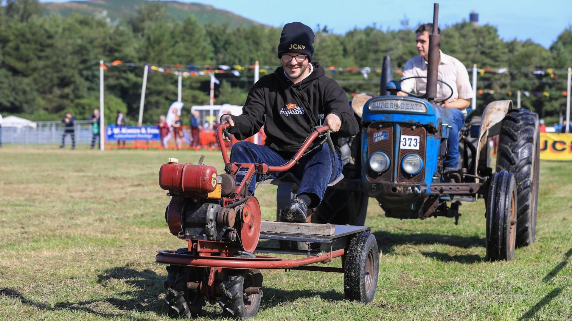 A man on a small very old tractor which is simply a seat on a platform with handle bars and wheels.