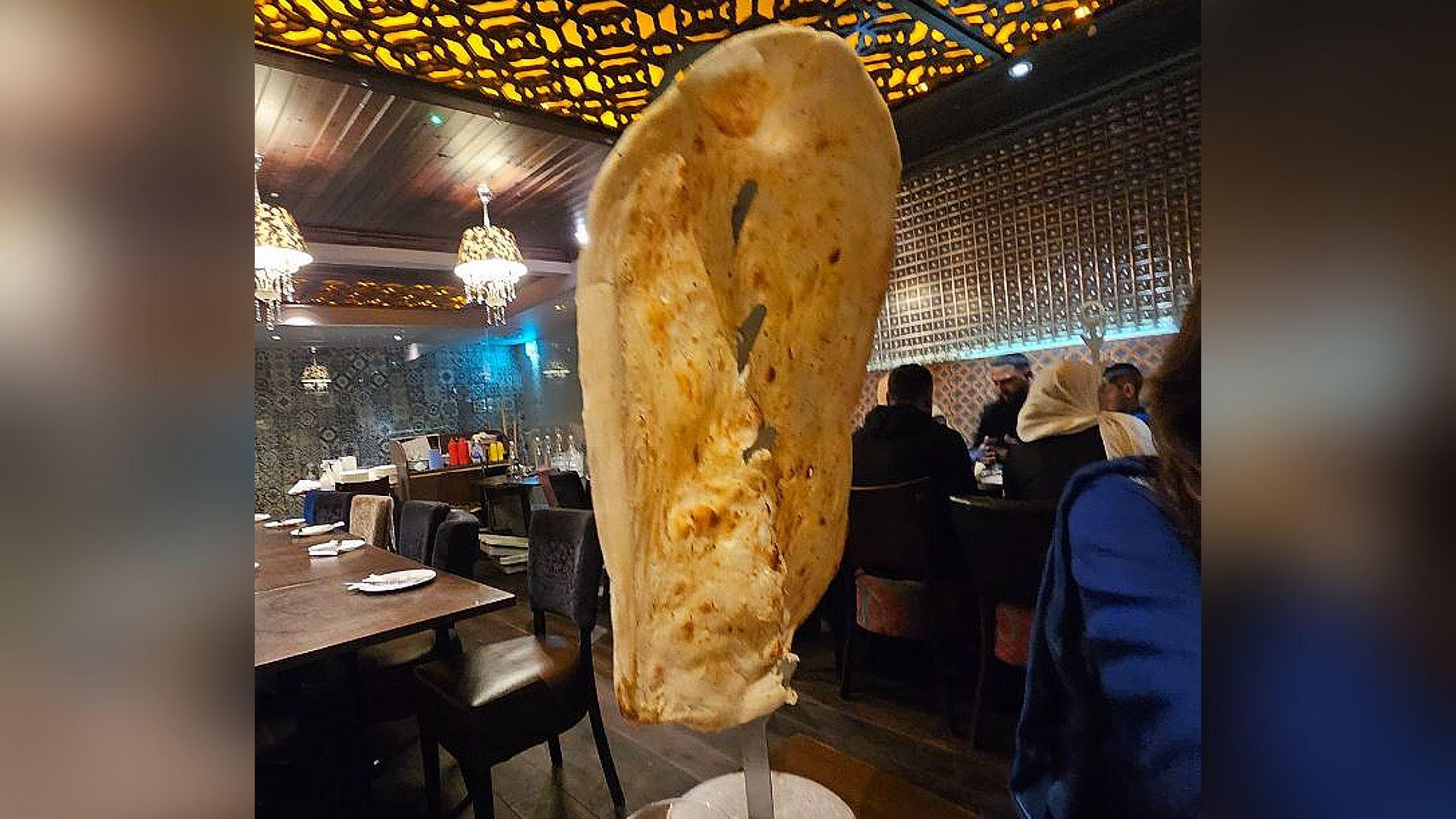 A large naan bread hangs on a vertical metal stand on a table in a restaurant setting. People are sat together eating on the right of the picture, with an empty table on the left side. 