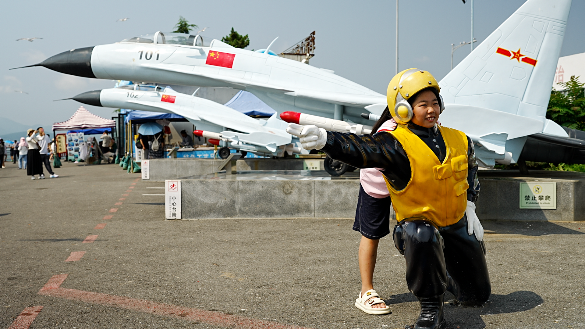 A girl in demin shorts and a pink shirt stand behind the statue of an airman. which is kneeling on the ground and pointing an arm in one direction- she puts her face above the statue's shoulders and smiles at the camera.
