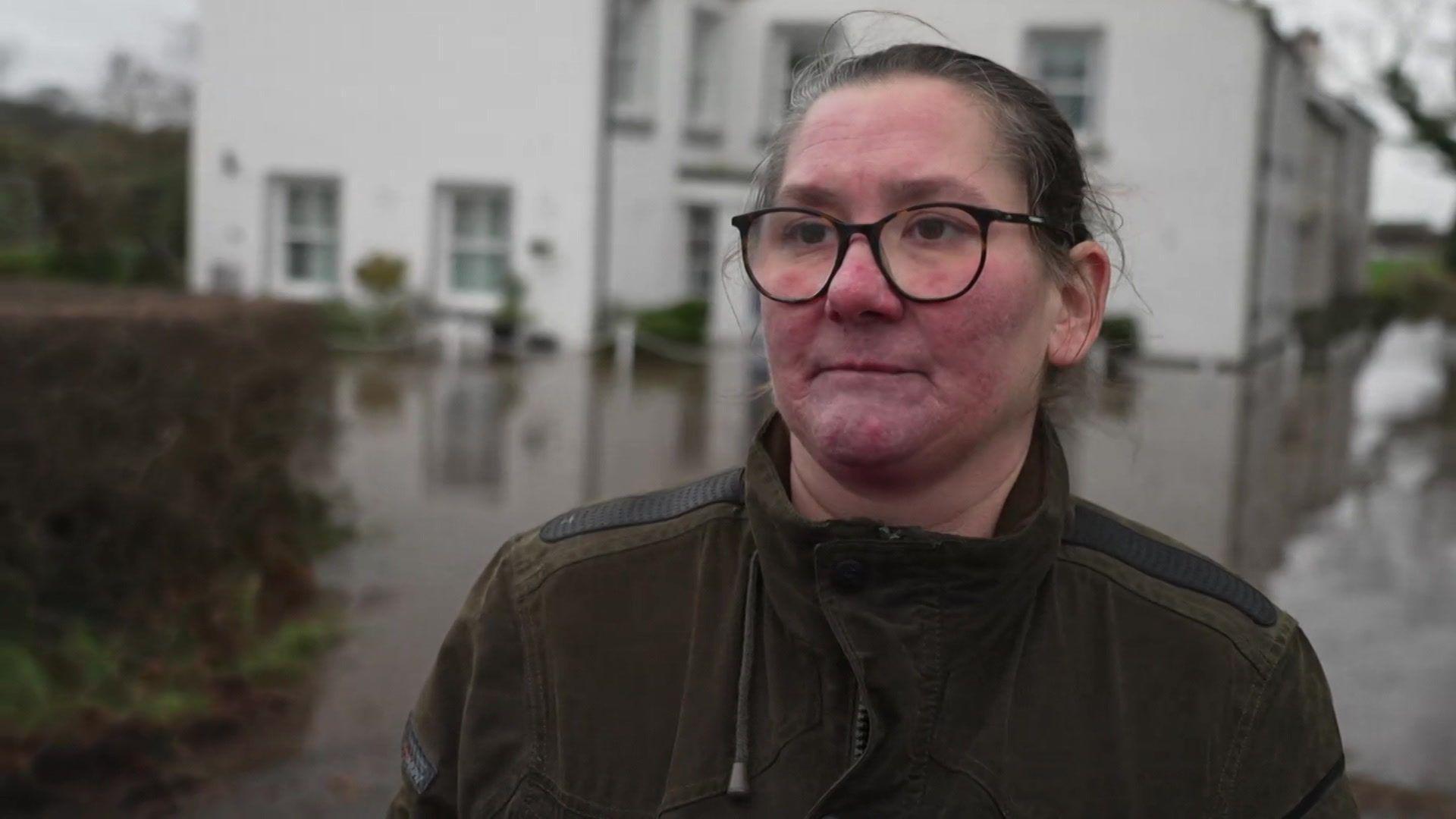 Gabrielle Dainty, who has black hair tied back and wears glasses and a green fleece, speaks to the camera with her flooded white cottage in the backdrop.