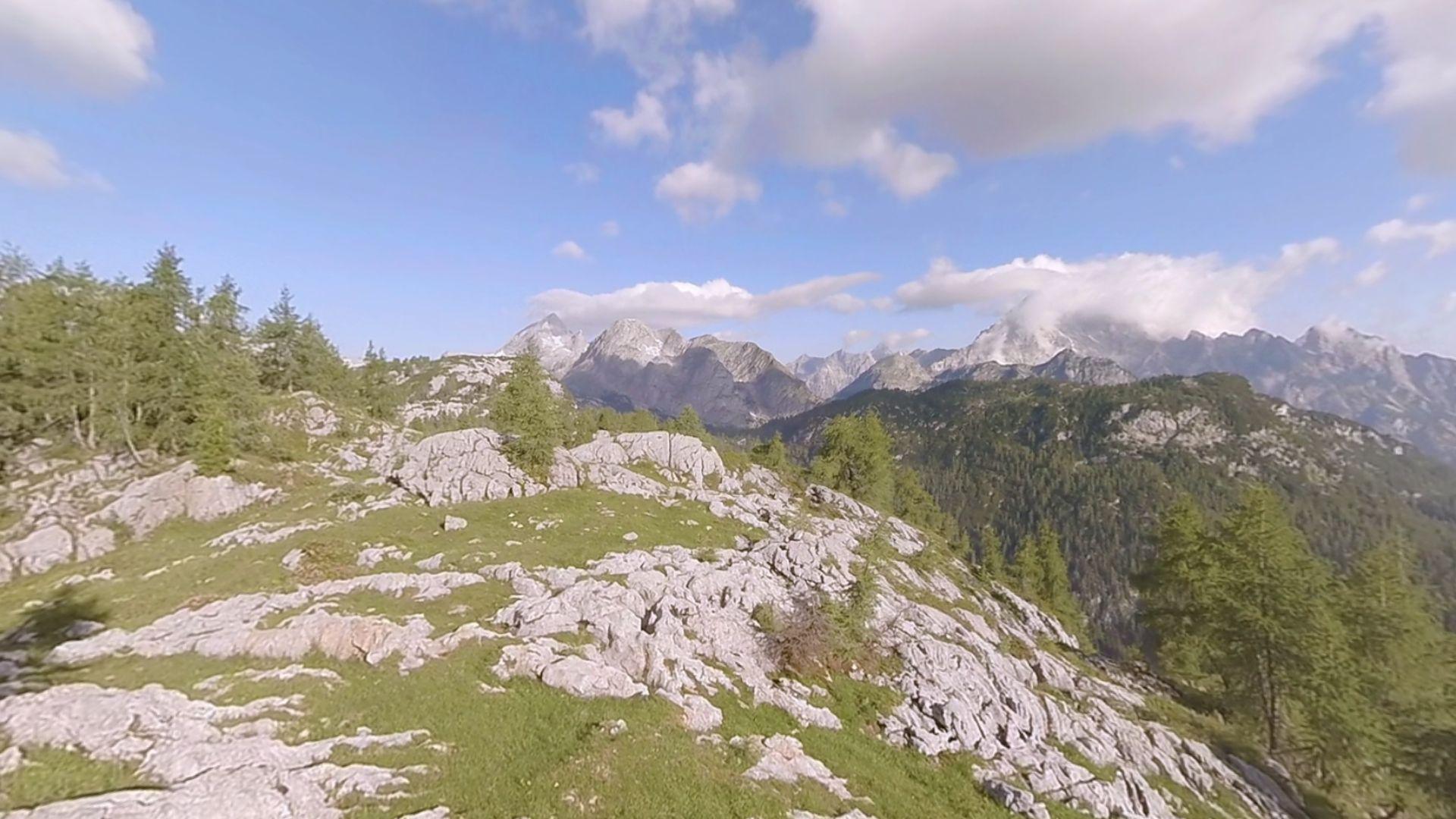 A mountain scene with grass and rocks on them. There are trees and a bright sky with a few clouds.