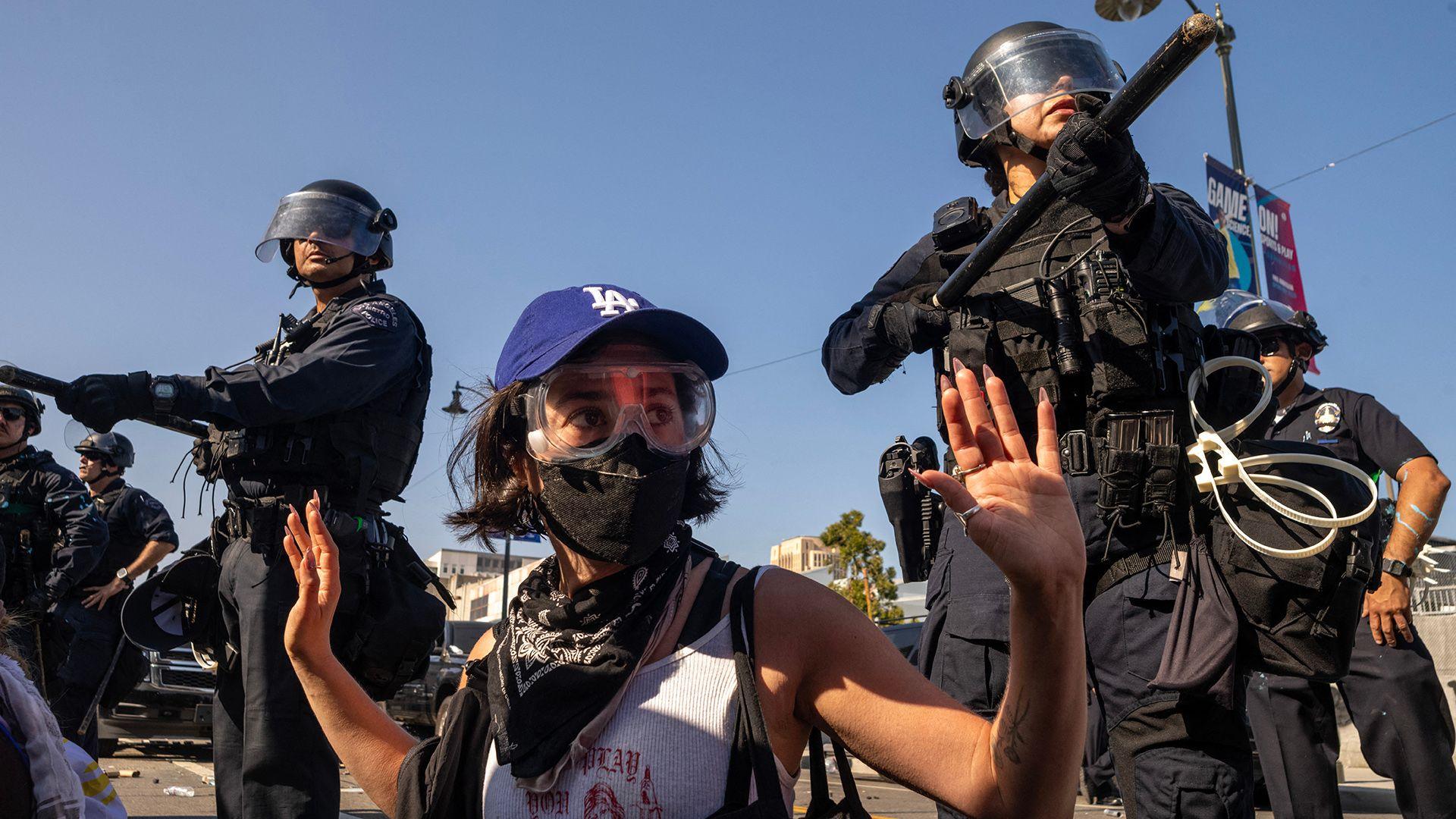 A protester wearing goggles and a face mask holds their hands in the air, while officers stand over them