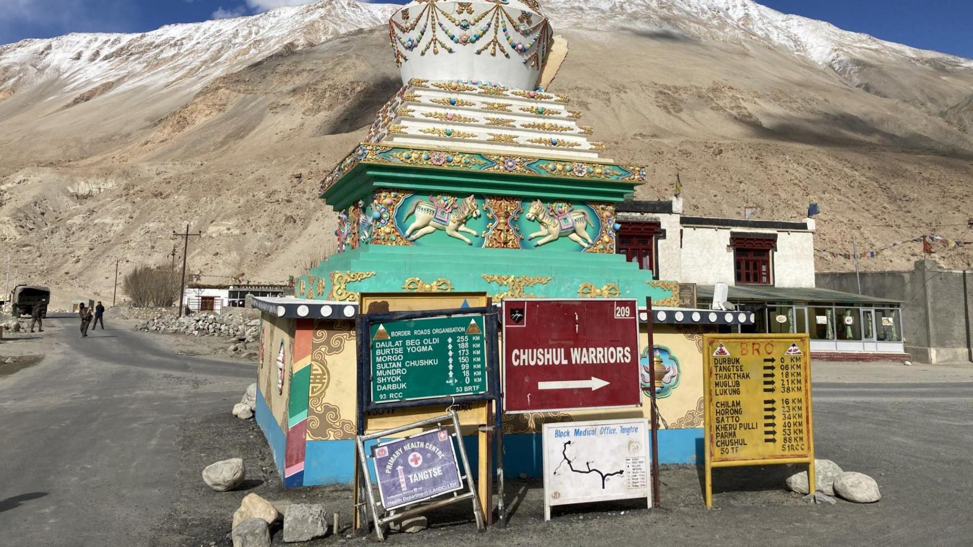 A view of the Chushul region in Ladakh with billboards in front of a decorative shrine, in the backdrop of stark, brown mountains.