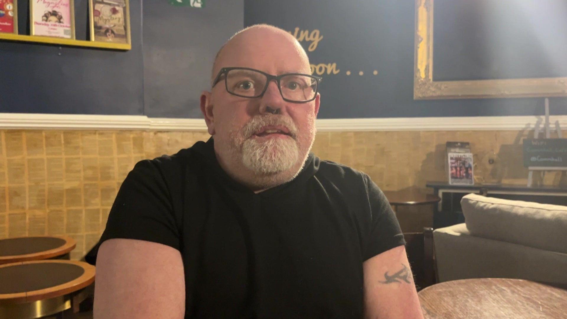 Michael Amps sitting in what looks like a cafe. He is bald and has a grey beard and moustache. He is wearing a black short-sleeve t-shirt and has dark square-framed glasses. He has a small Celtic tattoo on his upper right arm. 