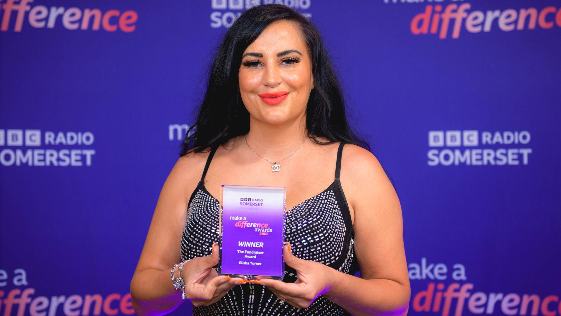 Woman with dark hair and red lipstick smiles at camera whilst holding a BBC Make a Difference trophy in front of her. The trophy is a small glass rectangle which is partly purple and partly white and has the details of her award in pale text on it