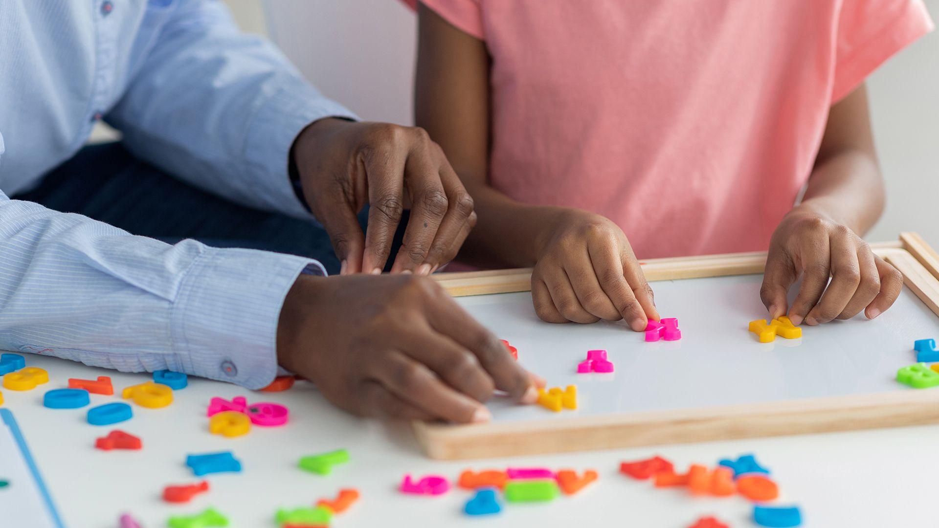 Stock photo shows a close up of the hands of a nursery teacher and young child arranging colourful letters onto a white desk while at nursery.