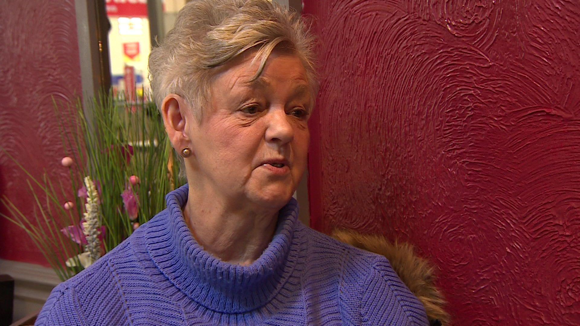 An elderly woman sits in a cafe wearing a lilac jumper