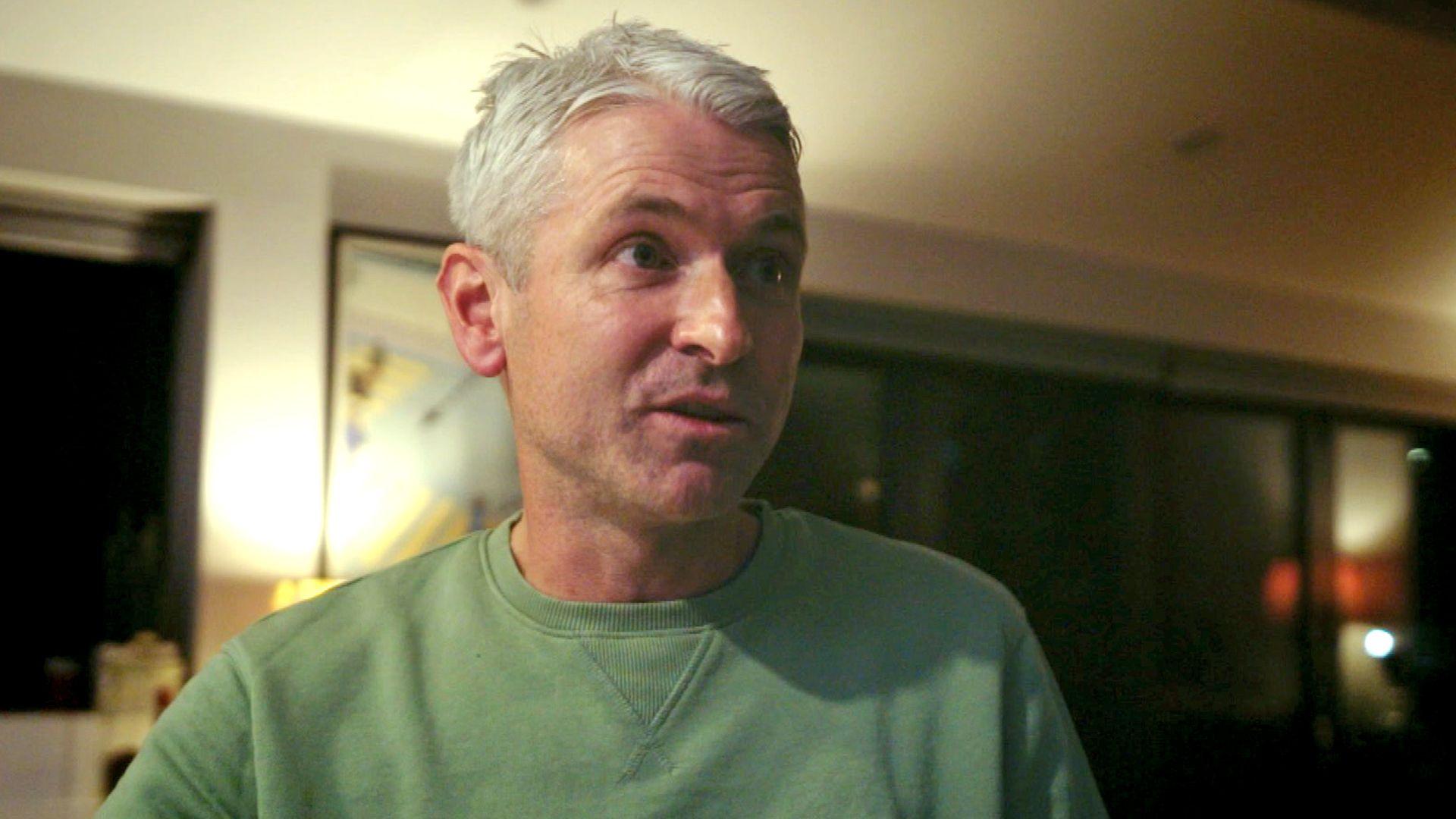 Rob Jones, with short grey hair wearing a green sweatshirt, stands indoors in a dimly lit room. The background features dark windows, a lit lamp, and framed artwork.