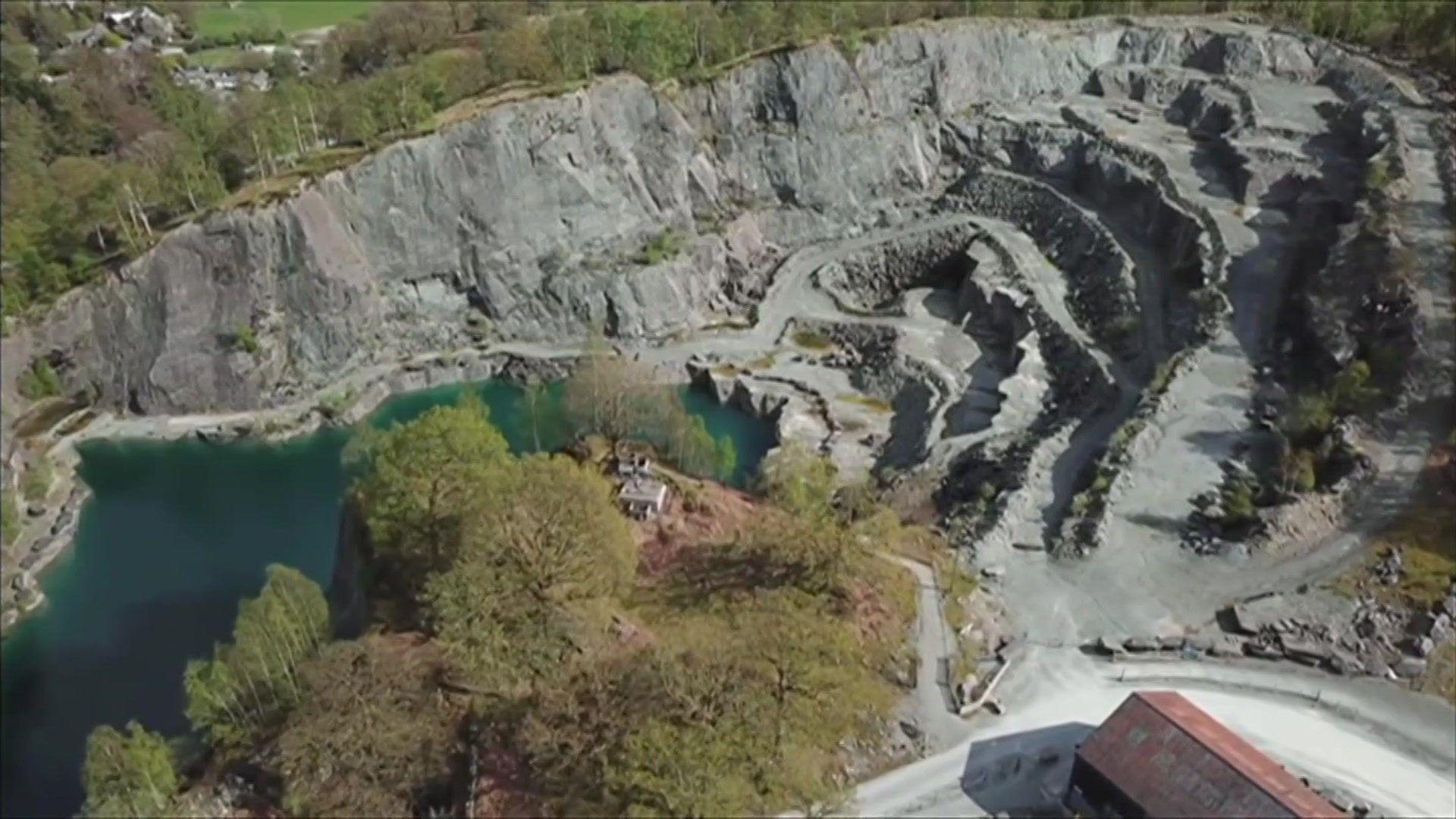 An aerial view of Elterwater Quarry. Layers of grey rock are to the right of a pool of water and some trees.