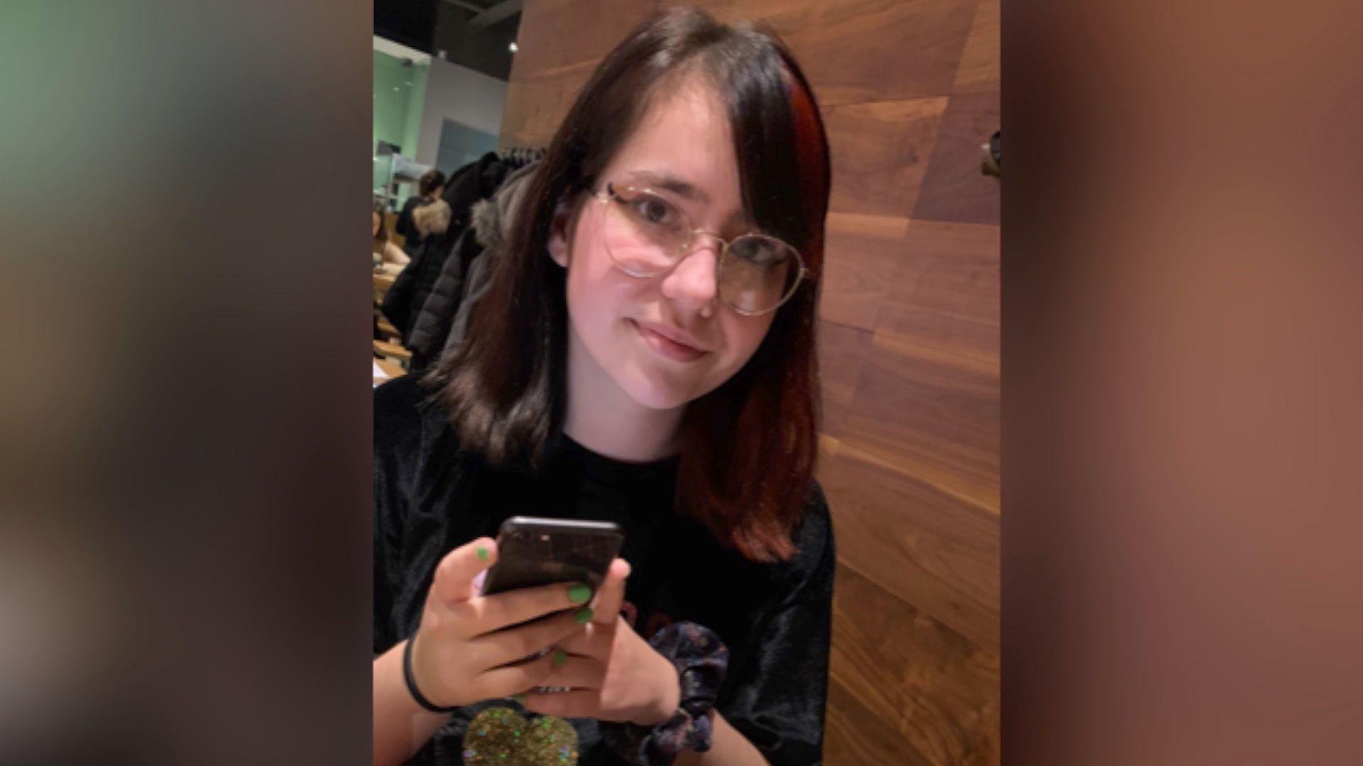 Elise Sebastian looking at a camera in a restaurant with a phone in her hand. She has green nail polish and is wearing metal rimmed glasses. She has brown hair with a red dyed tint to it.