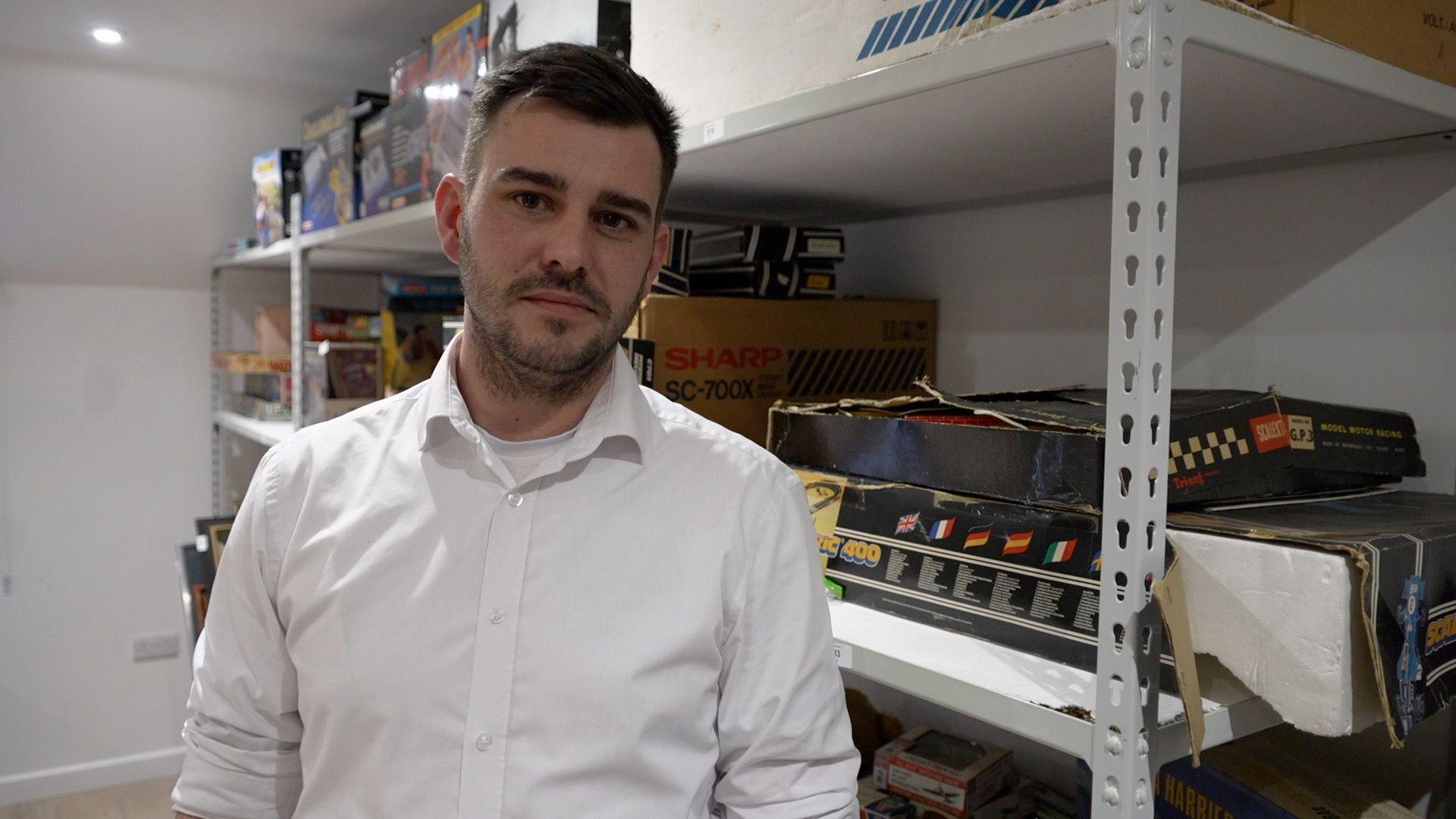 Daniel Pearce, in a white shirt, stands looking at the camera in the back room of his auction house. He has black hair and there are shelves behind him.